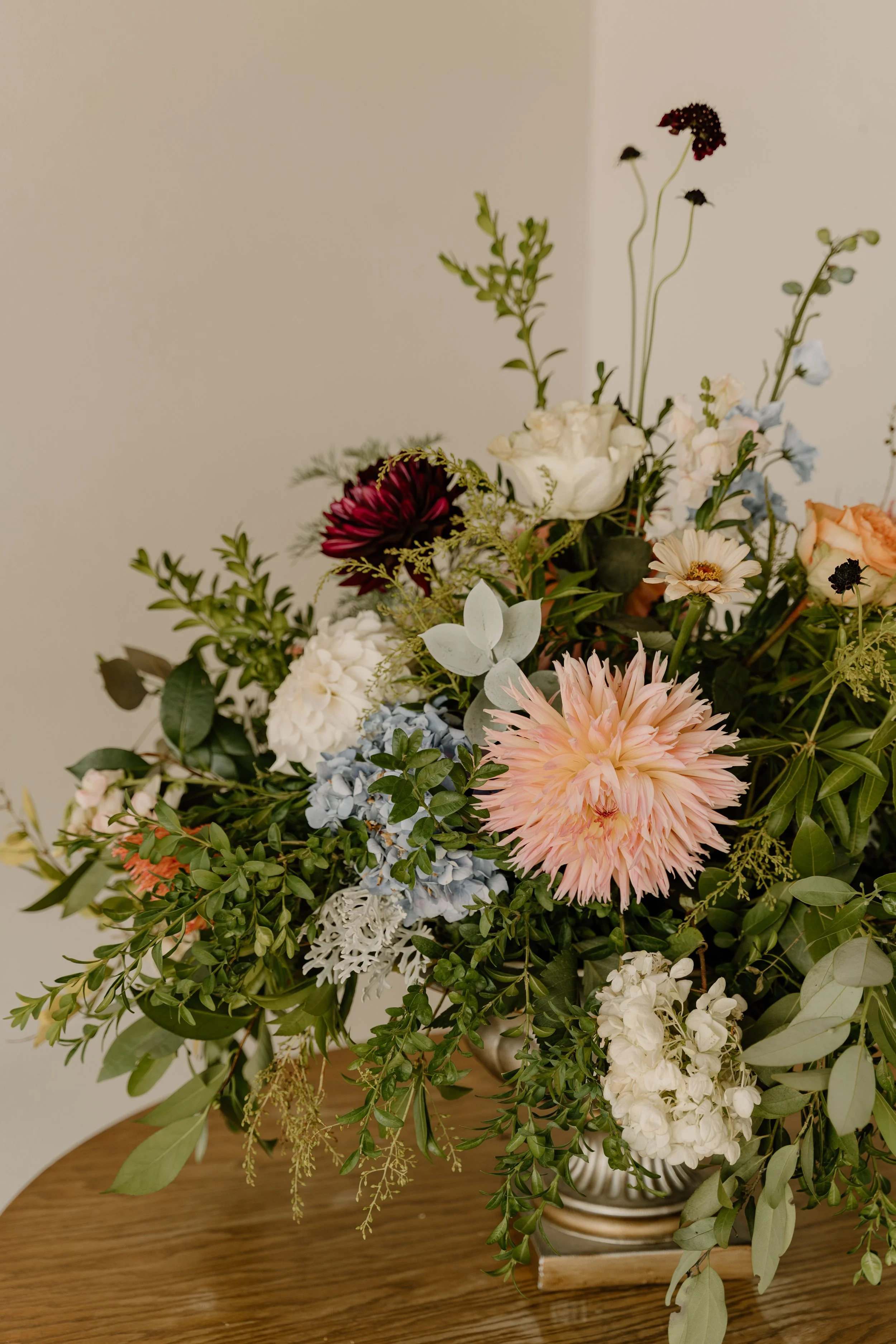 A floral arrangement with pink, white, and dark red flowers and greenery in a vase on a wooden table.