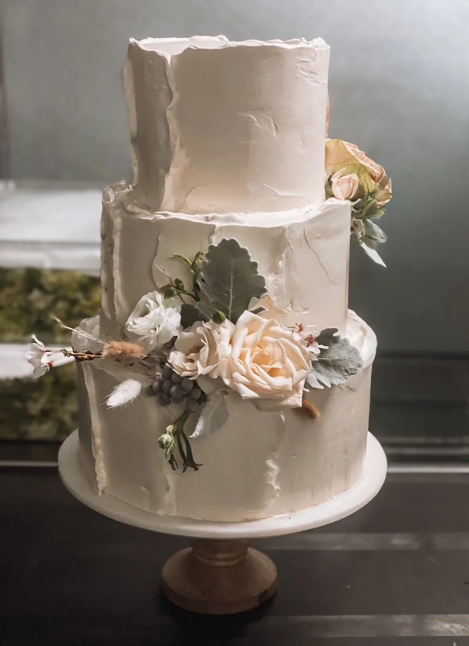 Three-tiered wedding cake with white frosting and decorative floral arrangements, including soft pink roses and green leaves.