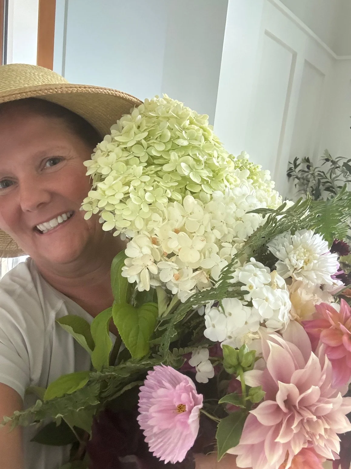 A smiling woman wearing a straw hat and white shirt holding a large bouquet of white, pink, and green flowers indoors.