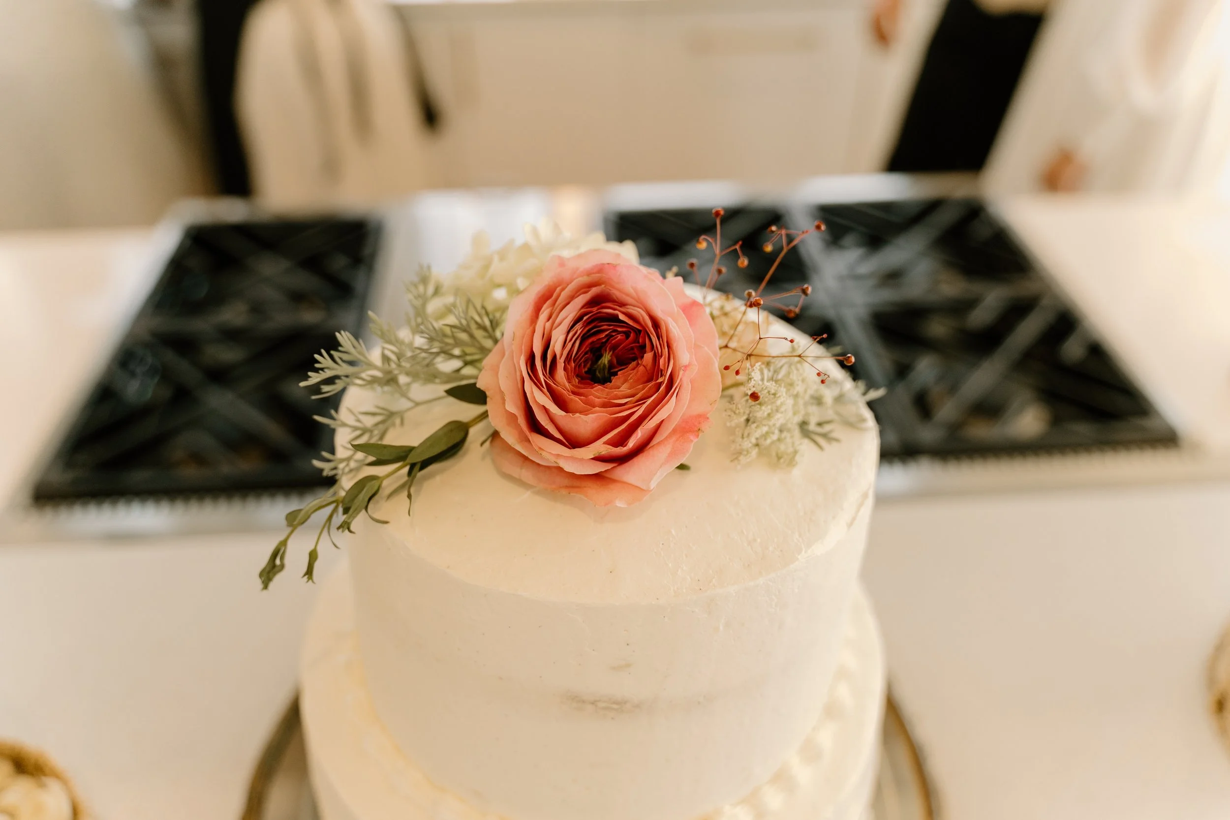 A two-tiered white wedding cake decorated with a pink ranunculus flower, green foliage, and small white and red accents on top. The cake is placed on a table with elegant black chairs and a black and white patterned table runner in the background.