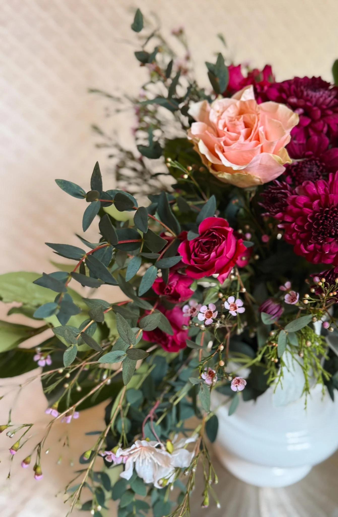 A floral arrangement with pink, red, and white flowers in a white vase, including roses, small white and pink blossoms, and green foliage.