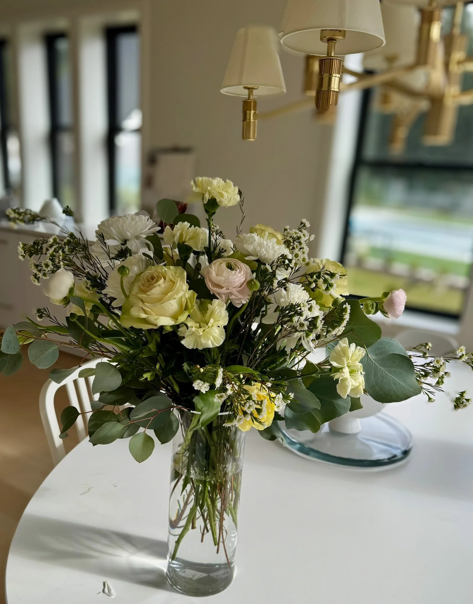 A glass vase filled with a bouquet of white and pale pink roses, lilies, and greenery on a white table in a bright room with large windows and a modern chandelier.