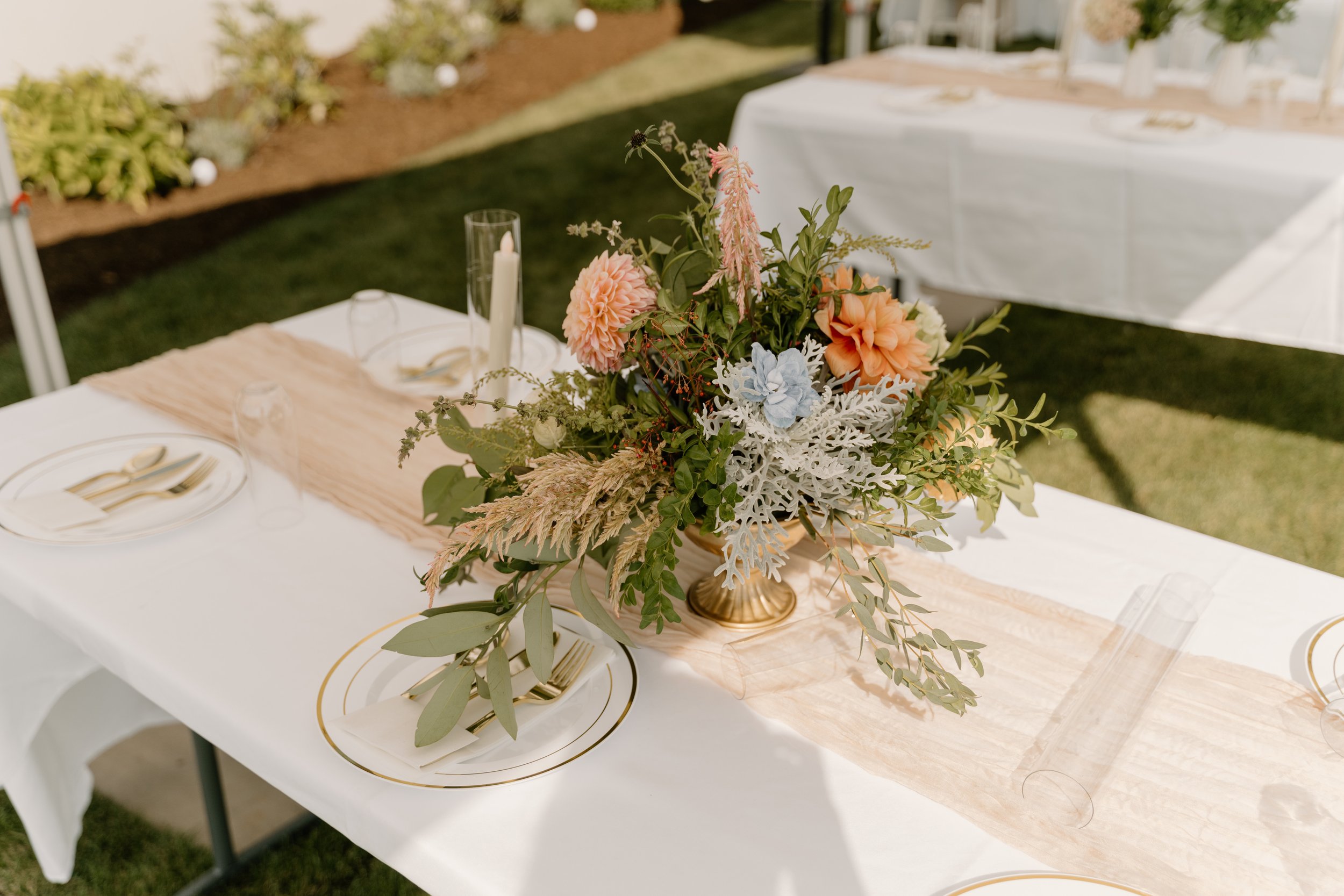 Elegant outdoor table setting with a large floral centerpiece in a gold vase, surrounded by white plates, gold utensils, a pink cloth table runner, and a white tablecloth, set on a grassy lawn.
