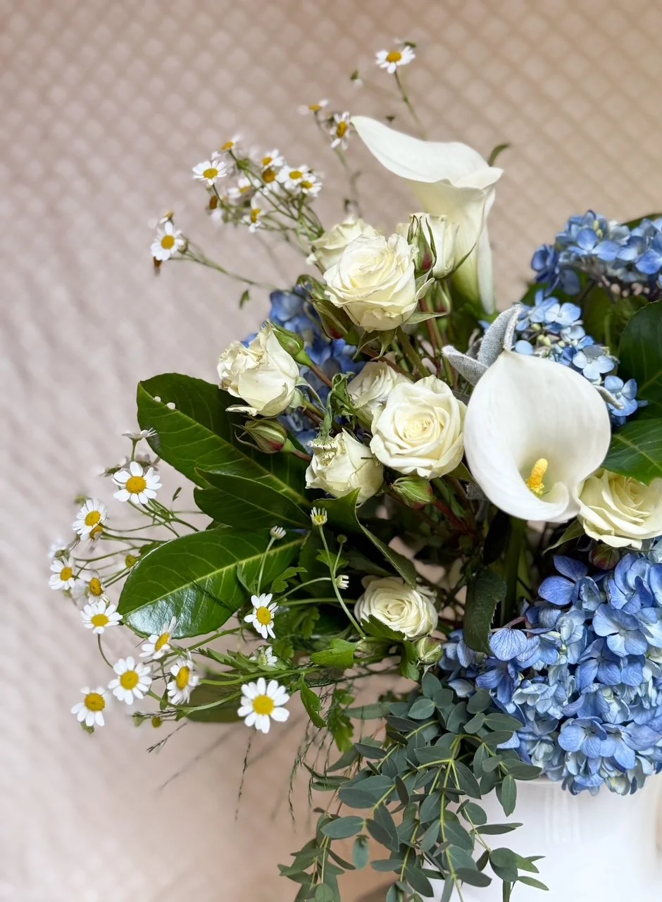 A floral arrangement with white roses, calla lilies, small white daisies, blue hydrangeas, and various green leaves in a white vase.