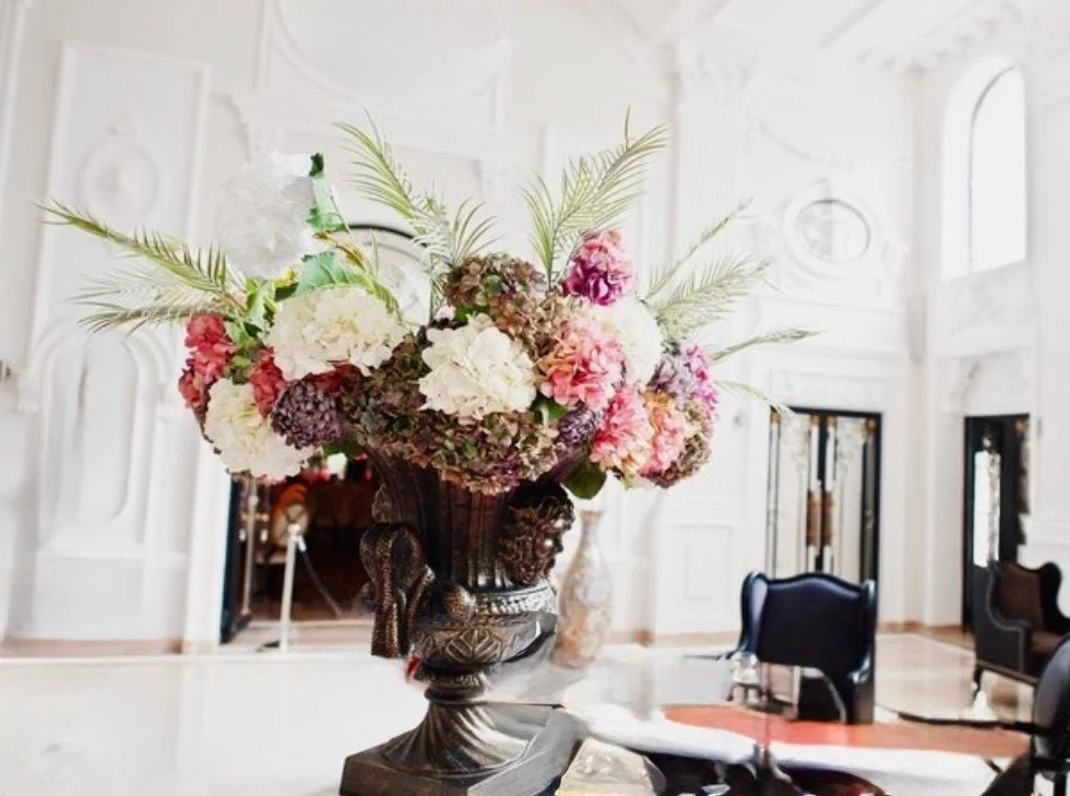 A large floral arrangement of white, pink, and purple hydrangeas with green leaves in a decorative silver vase on a marble table in a luxurious white room.