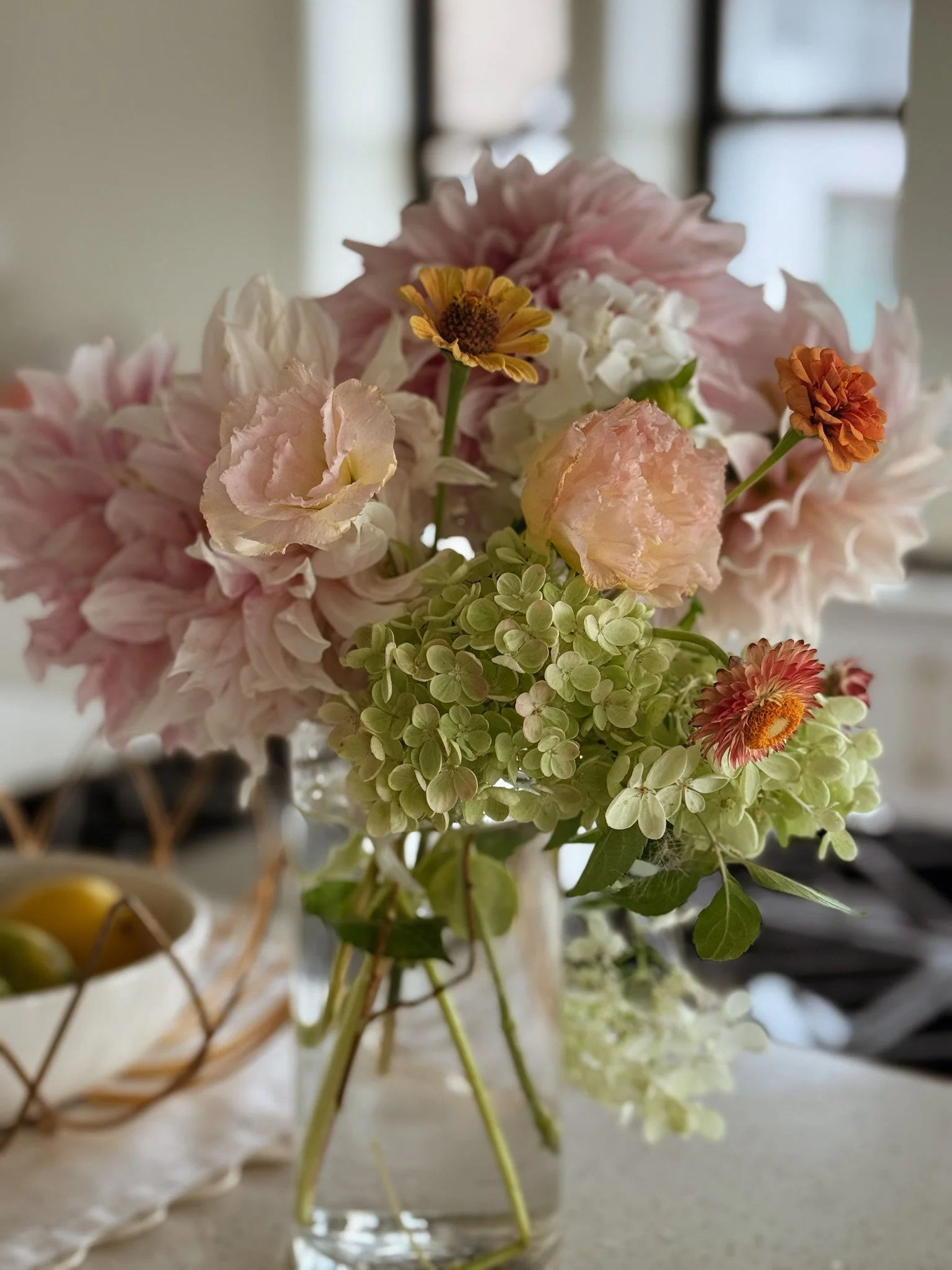 A glass vase with pink, white, and orange flowers, including hydrangeas, ranunculus, and daisies, on a table with a bowl of lemons in the background.