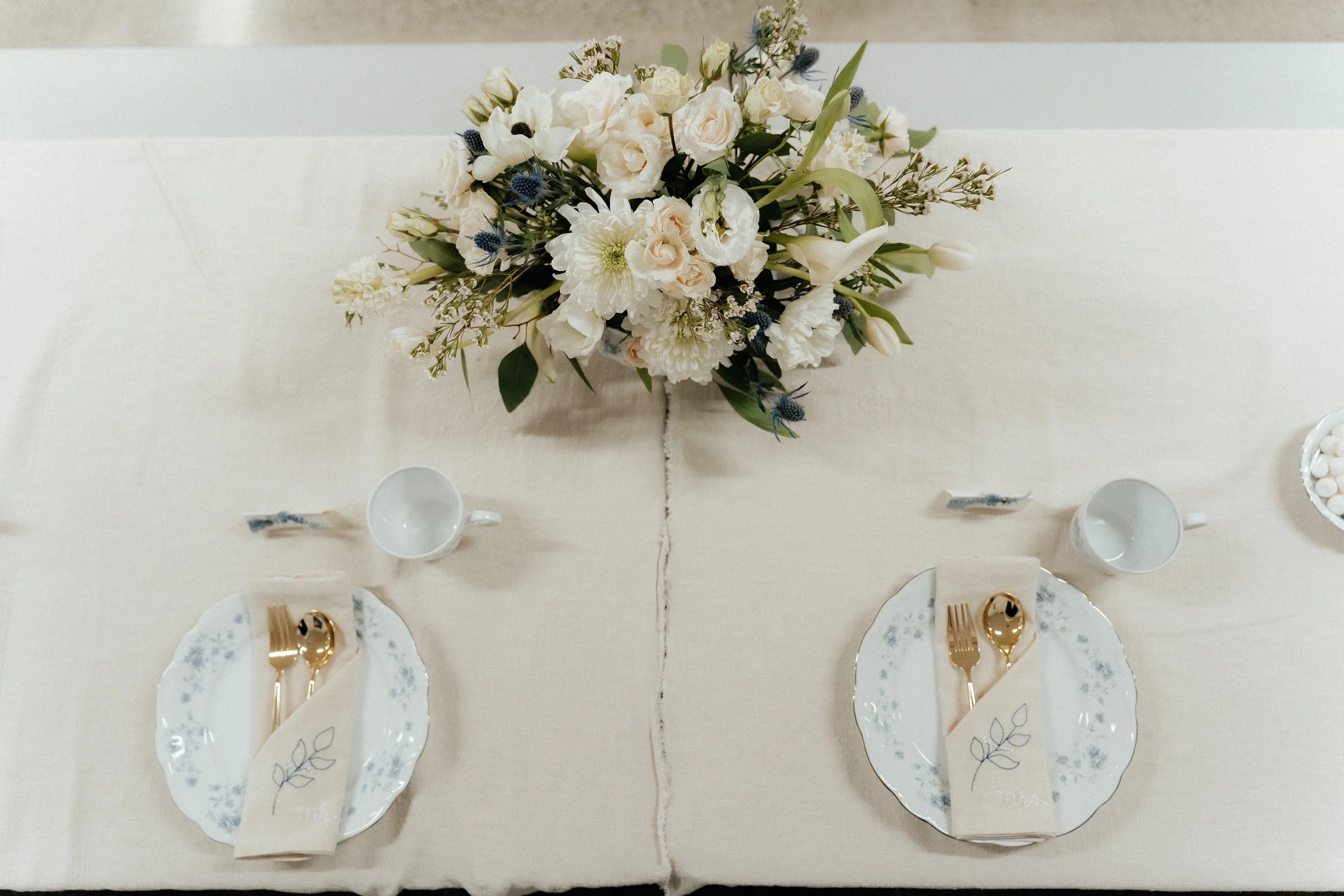 Table setting with a large floral centerpiece featuring white flowers, two place settings with white and blue patterned plates, gold utensils wrapped in napkins with leaf designs, and white cups.