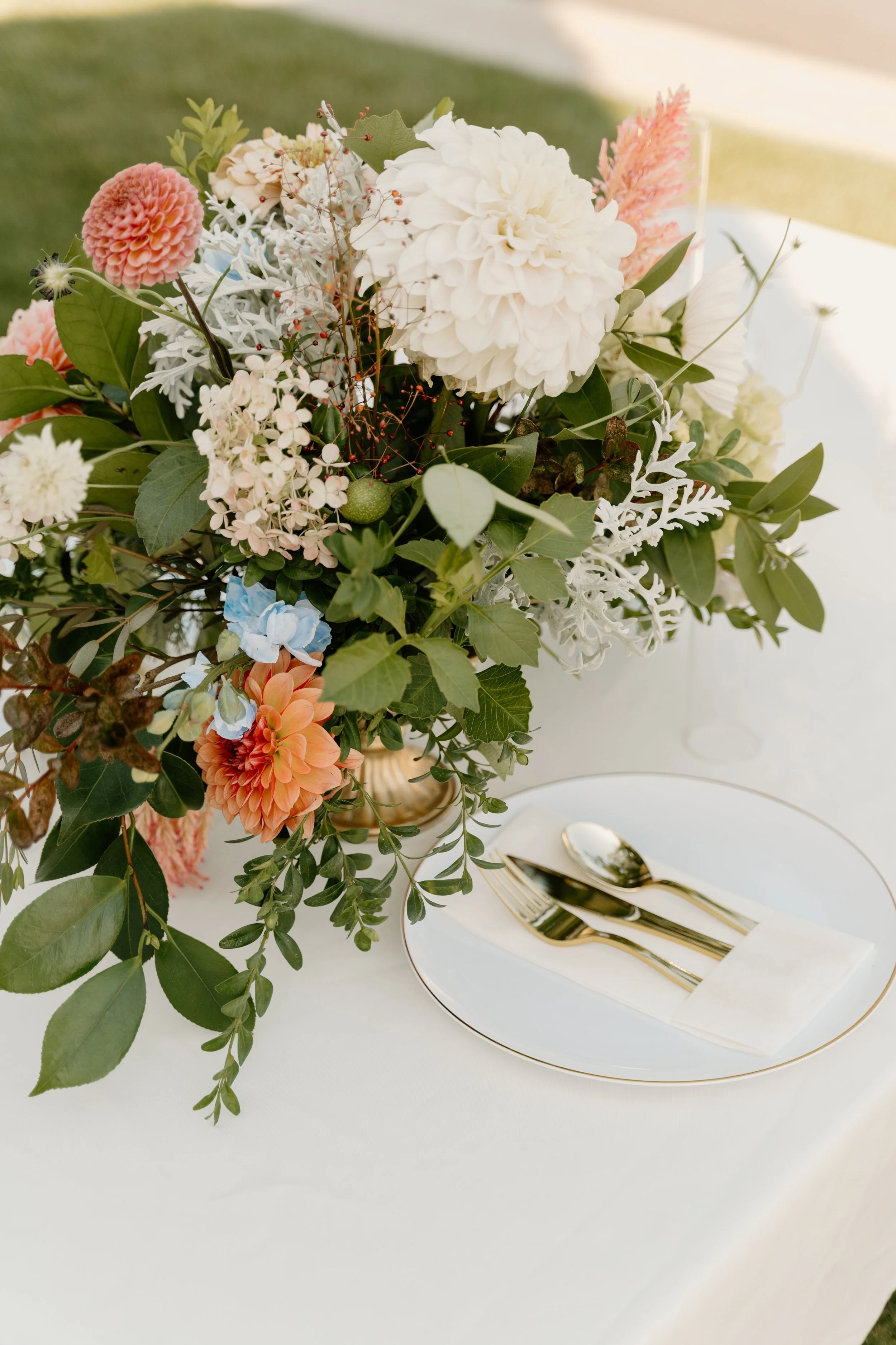 A floral centerpiece with white, pink, and peach-colored flowers and green leaves on a white table, with a set of gold cutlery on a white plate.