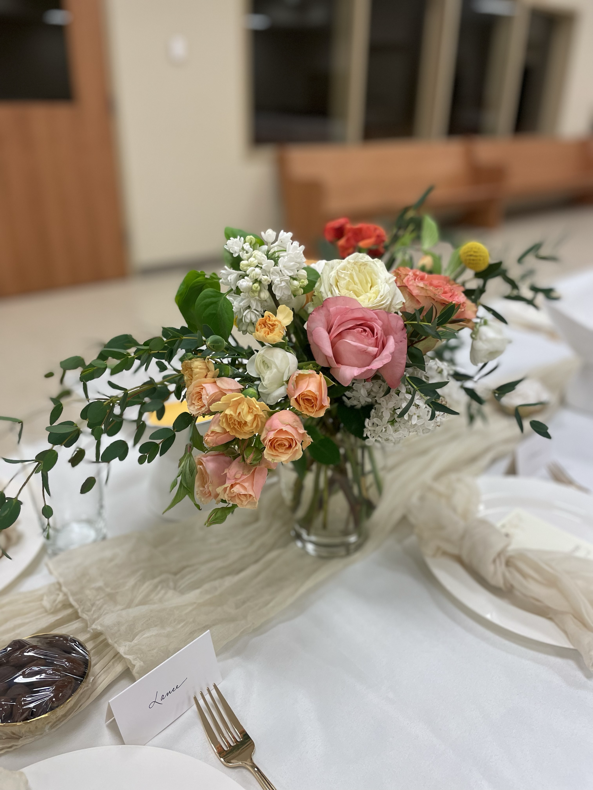 A floral centerpiece with pink, white, peach, and red flowers on a table with a white tablecloth and a cream-colored table runner, with place settings and a fork.
