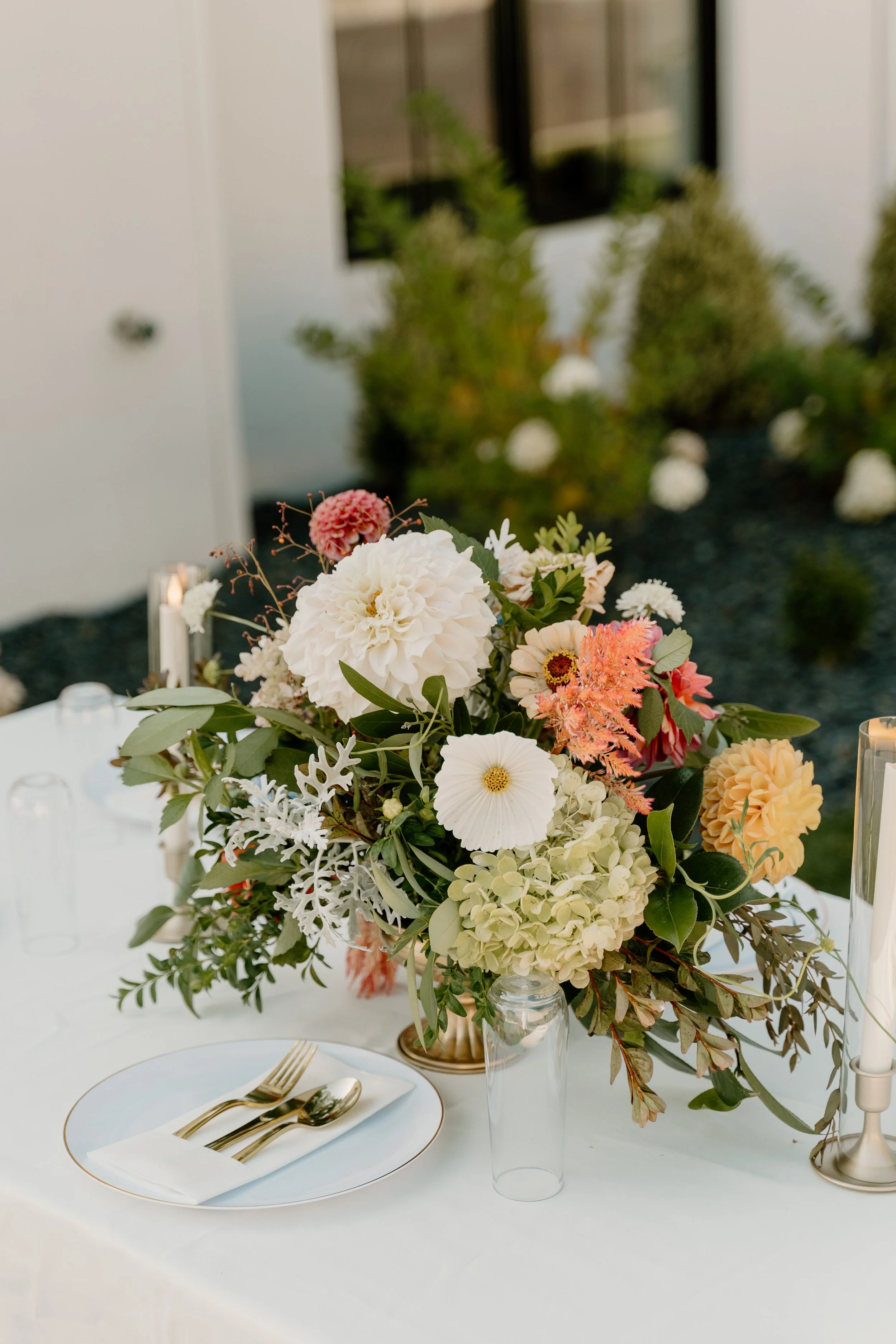 Elegant table centerpiece with pastel-colored flowers, including white dahlias, hydrangeas, and pink and peach blossoms, placed in a gold vase on a white tablecloth, with gold flatware and candles in the background.