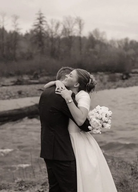A couple embracing outdoors near a river, with the woman holding a bouquet of flowers, both smiling.