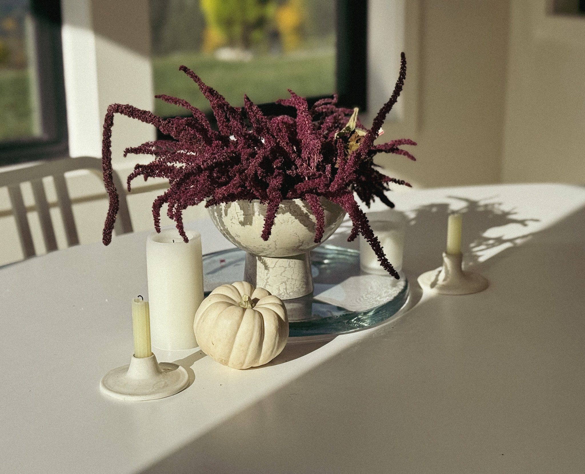 Autumn centerpiece on dining table with purple flowers in a white pot, a white pumpkin, and three white candles.