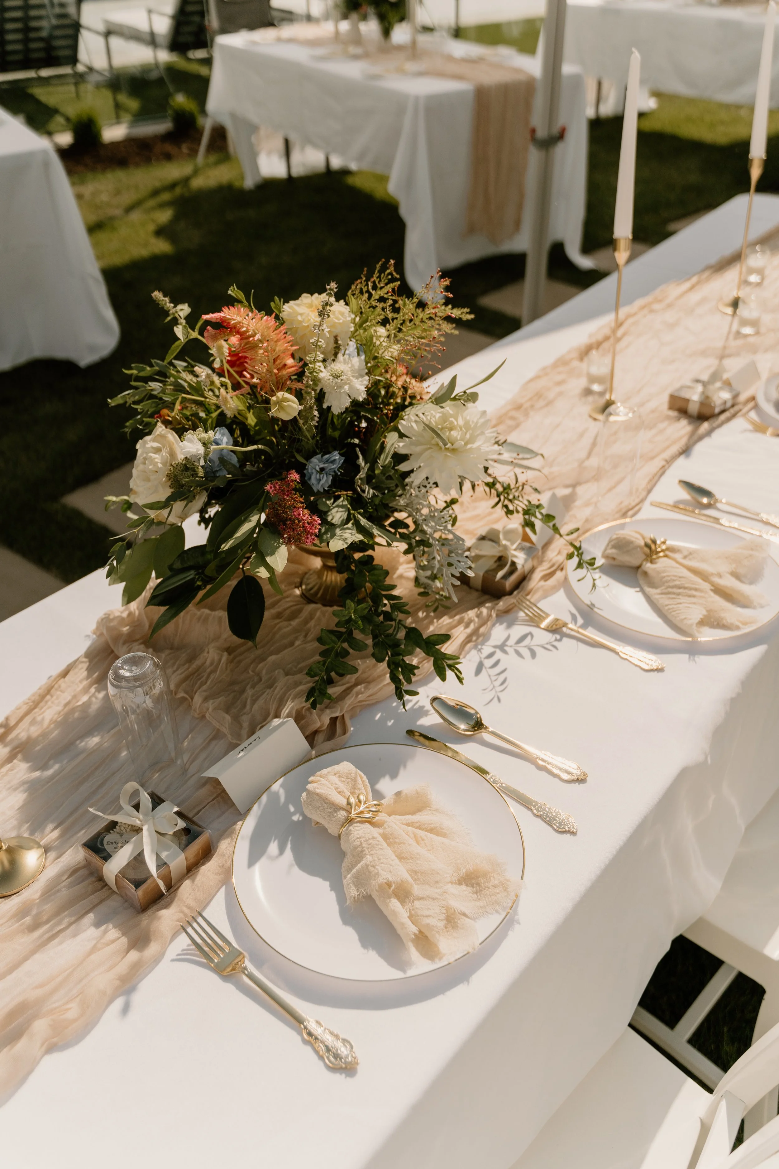 Elegant outdoor dining table set with white plates, gold flatware, cream napkins, a floral centerpiece, and candles with tall white candles. The table features a beige table runner and is surrounded by white chairs.