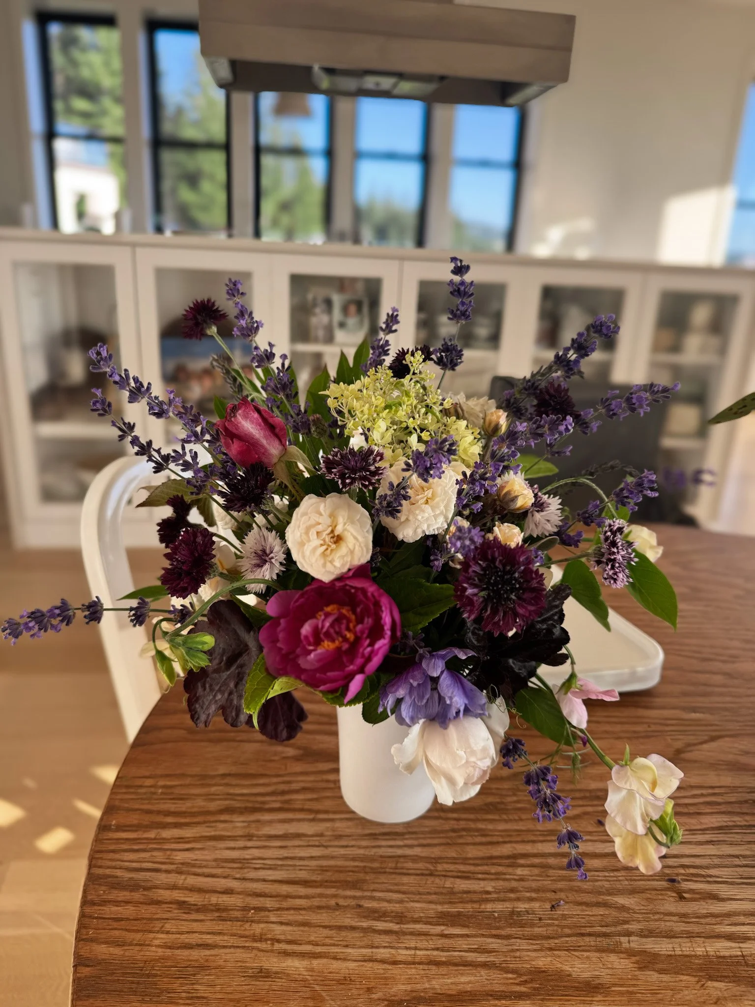 A colorful bouquet of flowers in a white vase on a wooden table, with a background of a room with large windows and a white cabinet filled with photos and items.