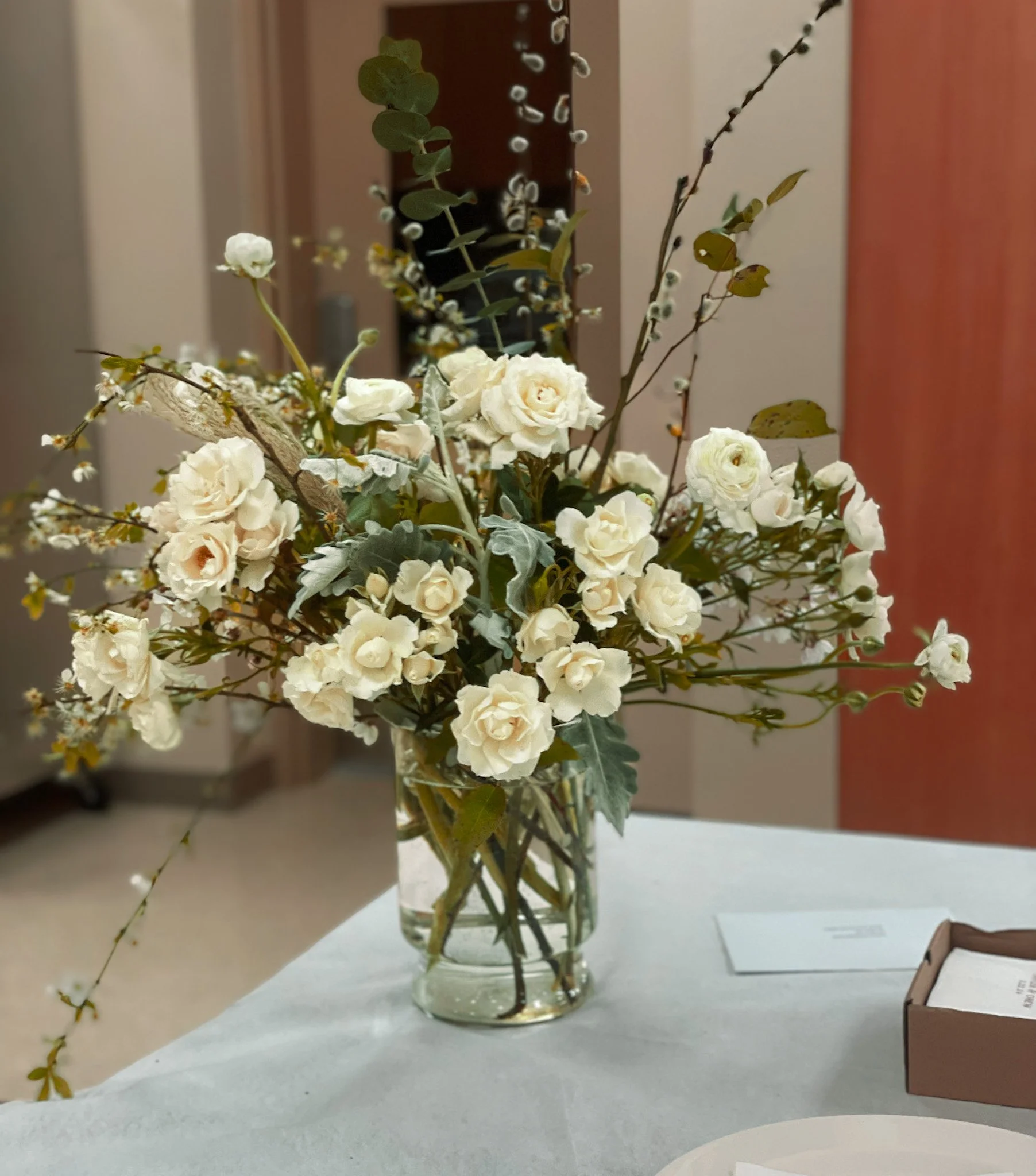 A glass vase filled with white roses, green foliage, and decorative branches sitting on a table.