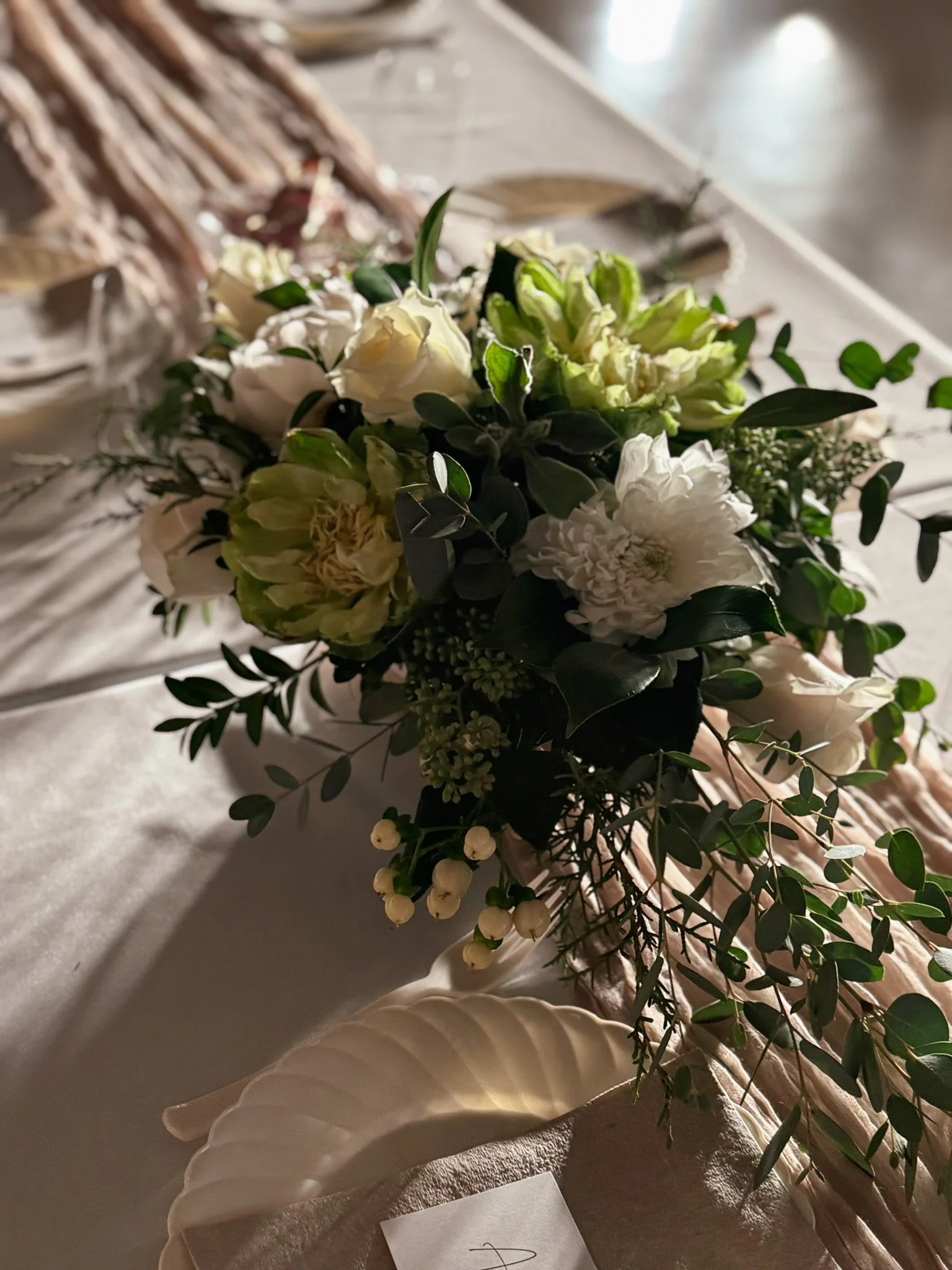 A floral centerpiece with white and green flowers and greenery on a table, with a white shell-shaped plate nearby.