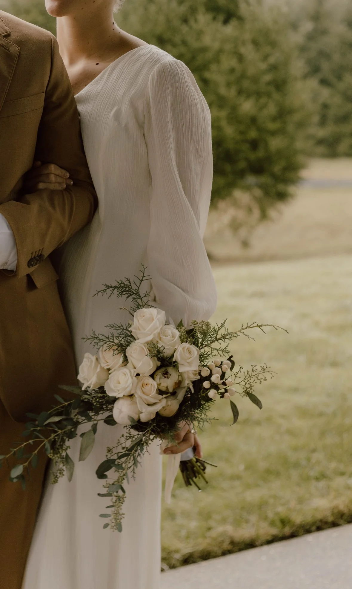A bride holding a bouquet of white roses and greenery, standing next to a groom in a tan suit outdoors.