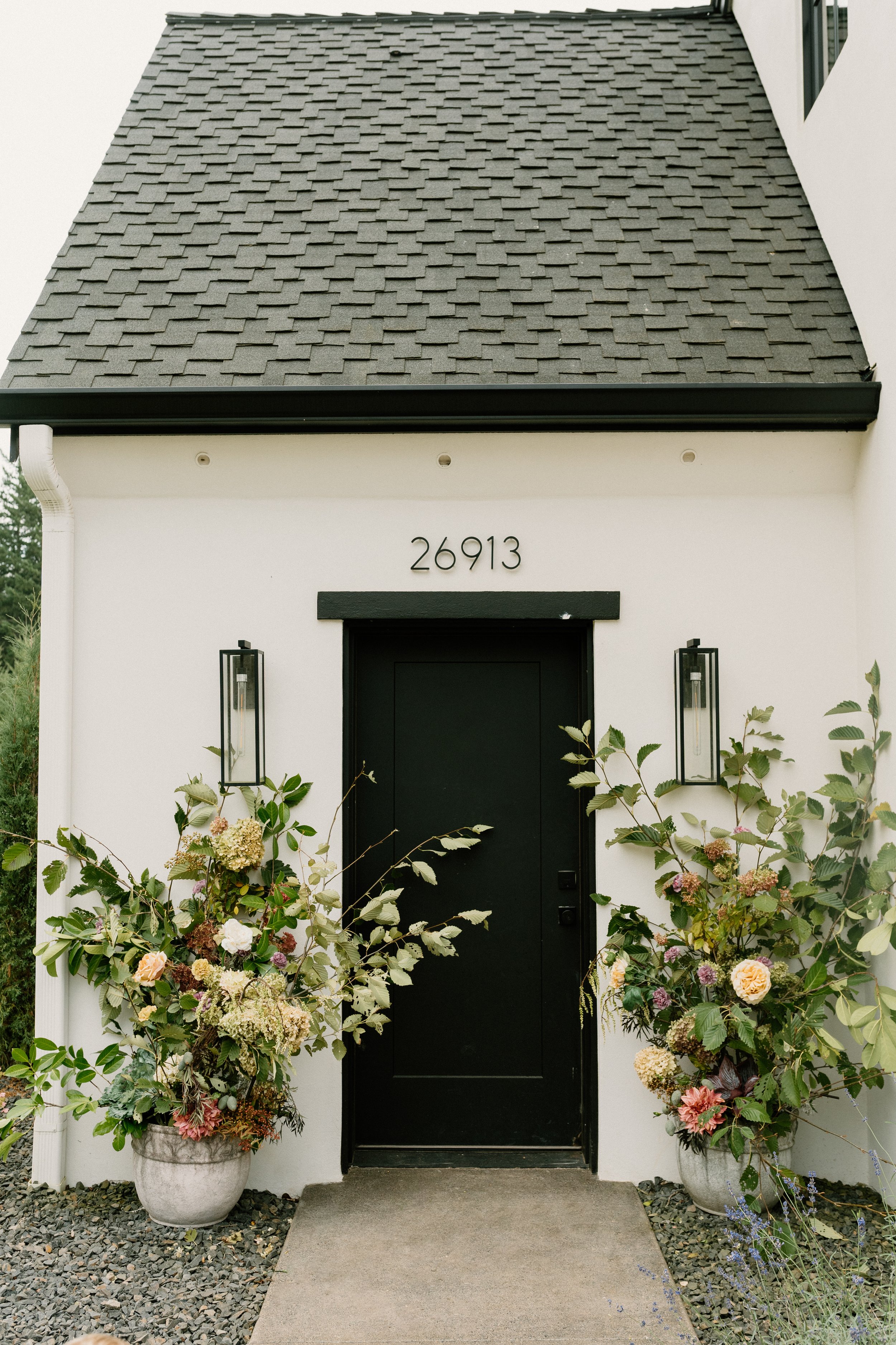Front entrance of a house with black door, white walls, and decorative flower arrangements in large vases on either side of the door, with house number 26913 above the door.