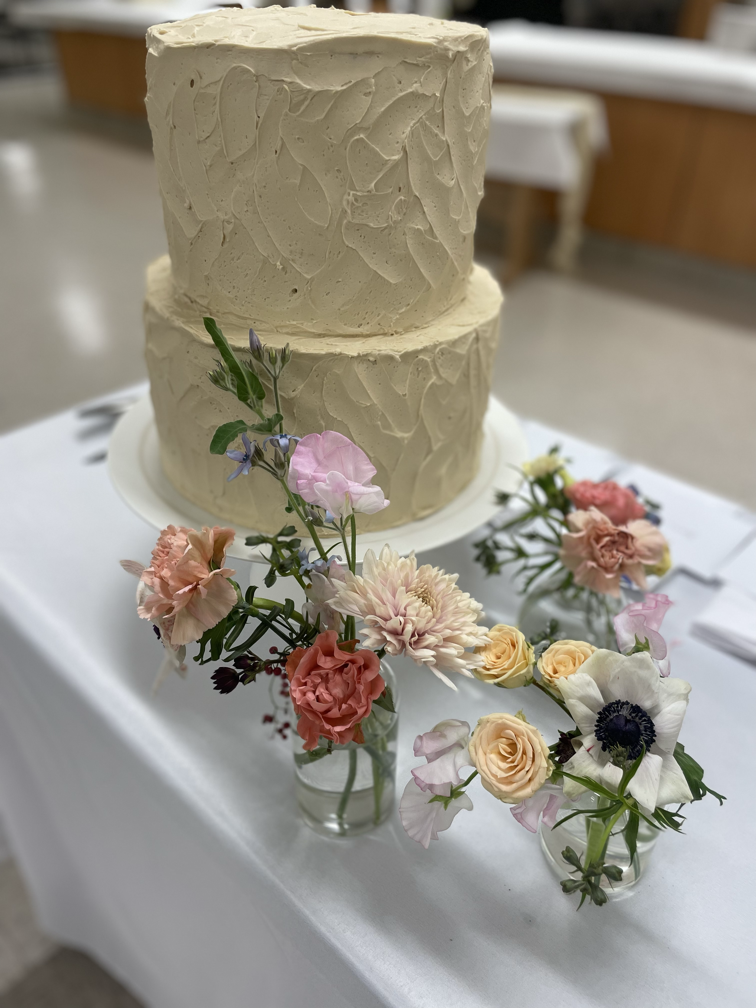 A double-tiered wedding cake with beige textured frosting, decorated with a bouquet of pink, peach, white, and purple flowers in front of it. The cake is on a white tablecloth with some silverware and paper visible nearby.