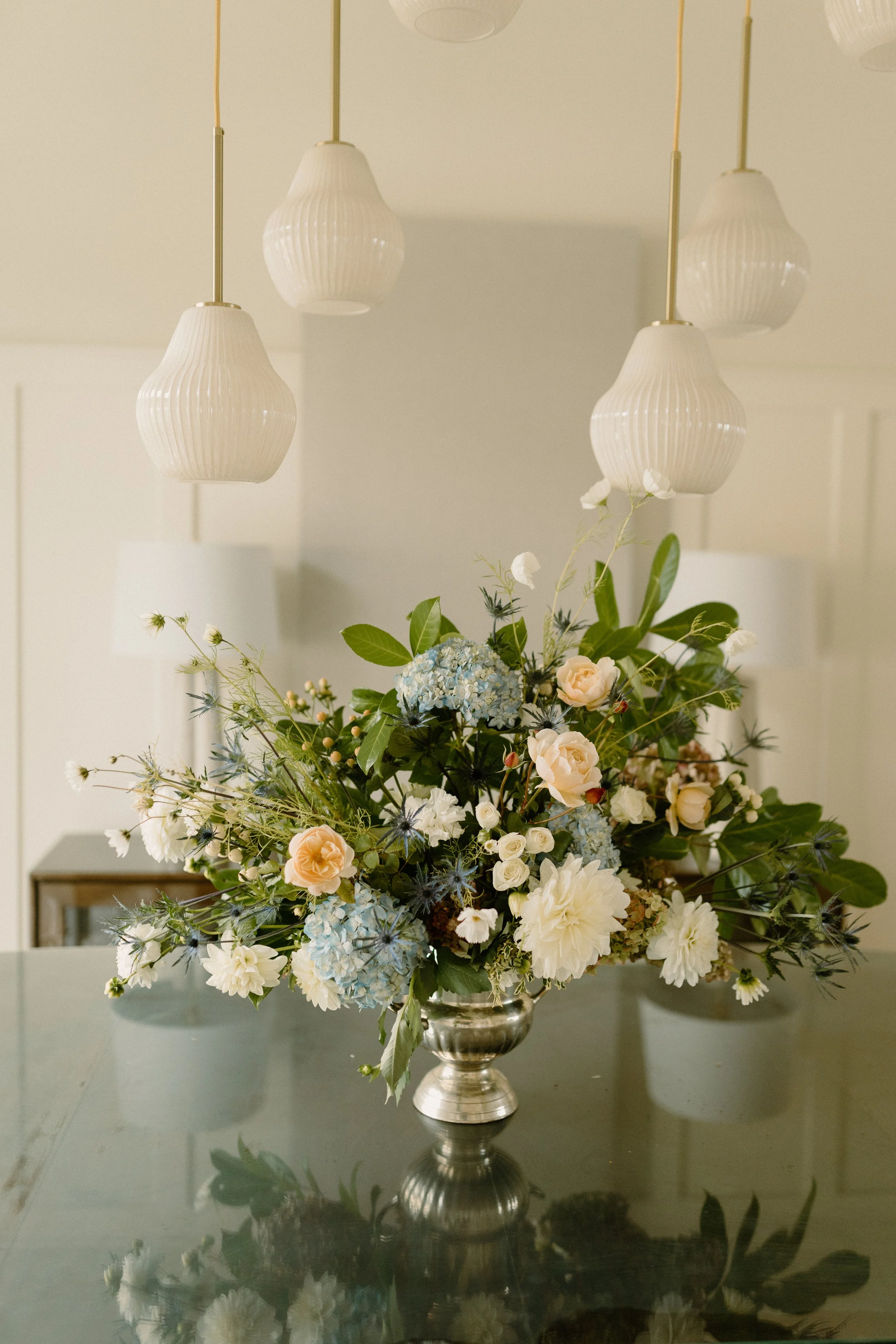 A floral arrangement with white, blue, and peach flowers in a silver vase on a reflective table.