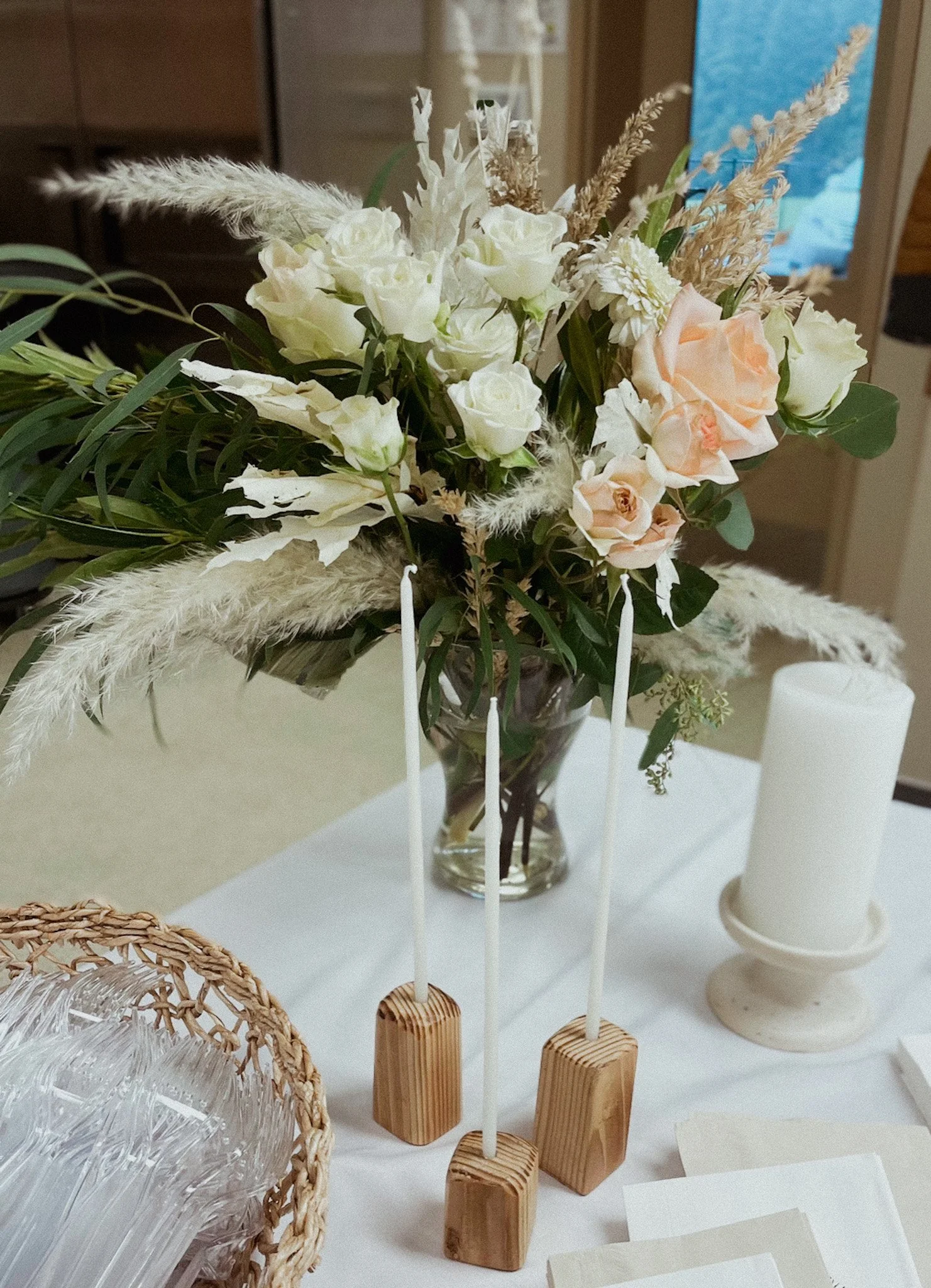 A floral arrangement in a glass vase with white and soft pink roses, greenery, and dried grasses, placed on a table beside white candles and a woven basket.
