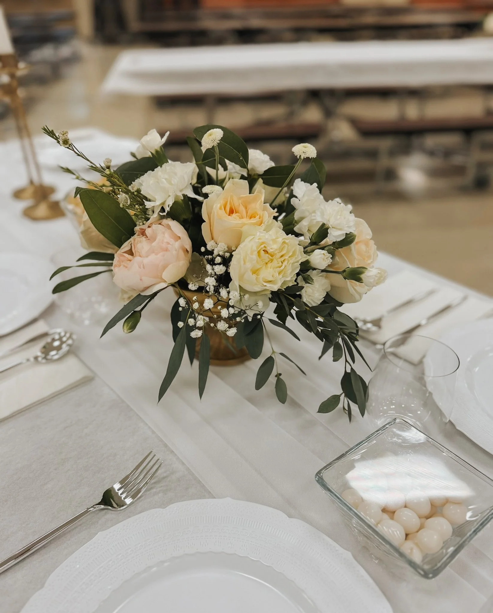 A table centerpiece with white and cream roses, peonies, and greenery. The table is set with white plates, silverware, and a glass bowl filled with white candies or mints.
