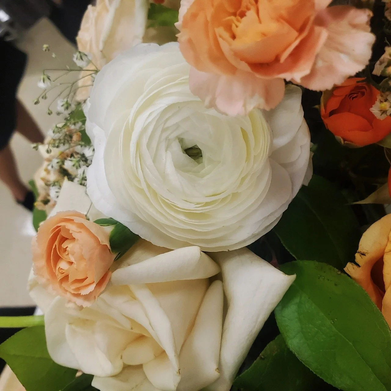 Close-up of a bouquet of flowers featuring white ranunculus, light pink roses, orange roses, and greenery.
