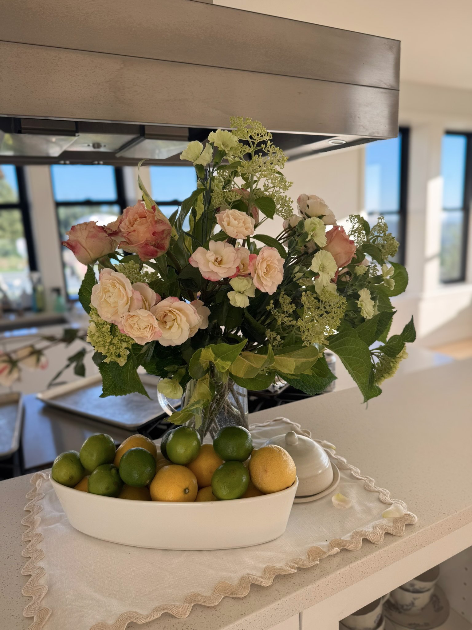 A kitchen countertop with a vase of pink and white flowers, a white bowl of green and yellow lemons, and a small white container on a lace-edged cloth in a bright room with large windows.