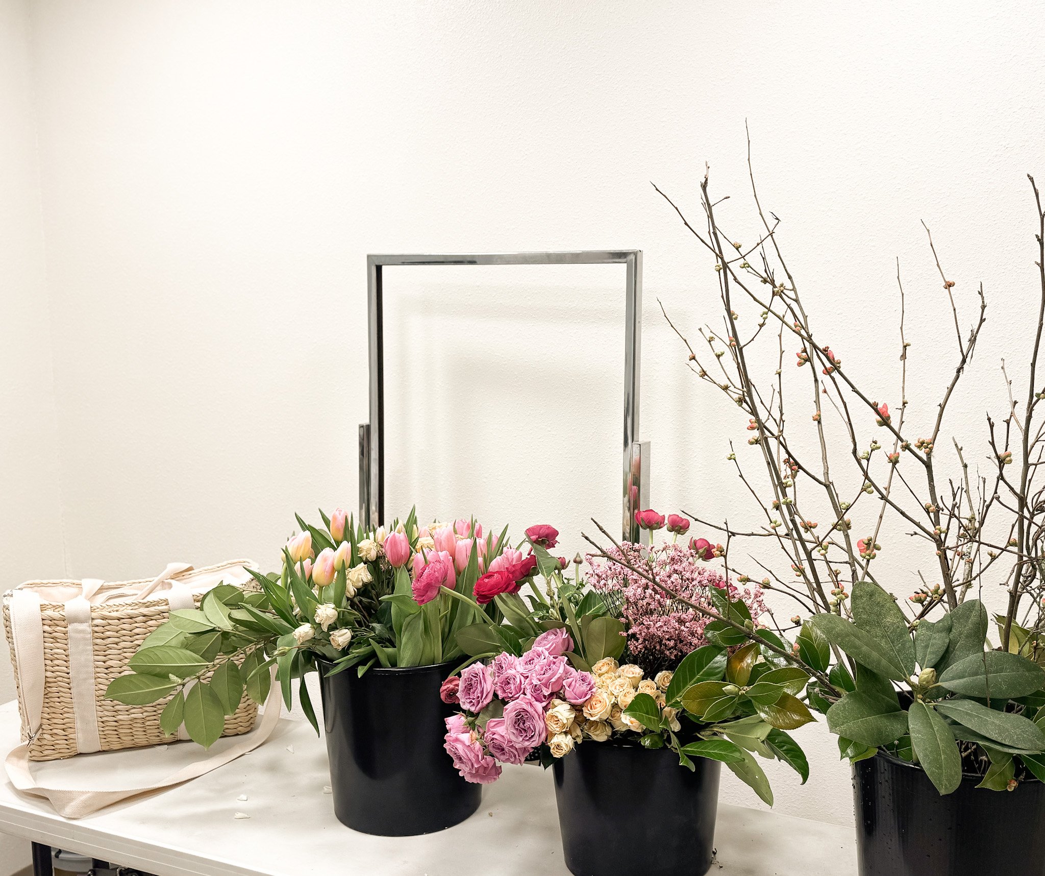 Various flower arrangements with pink tulips, roses, and other blooms in black pots, a straw basket, and bare branches with budding flowers on a white table against a plain white wall.