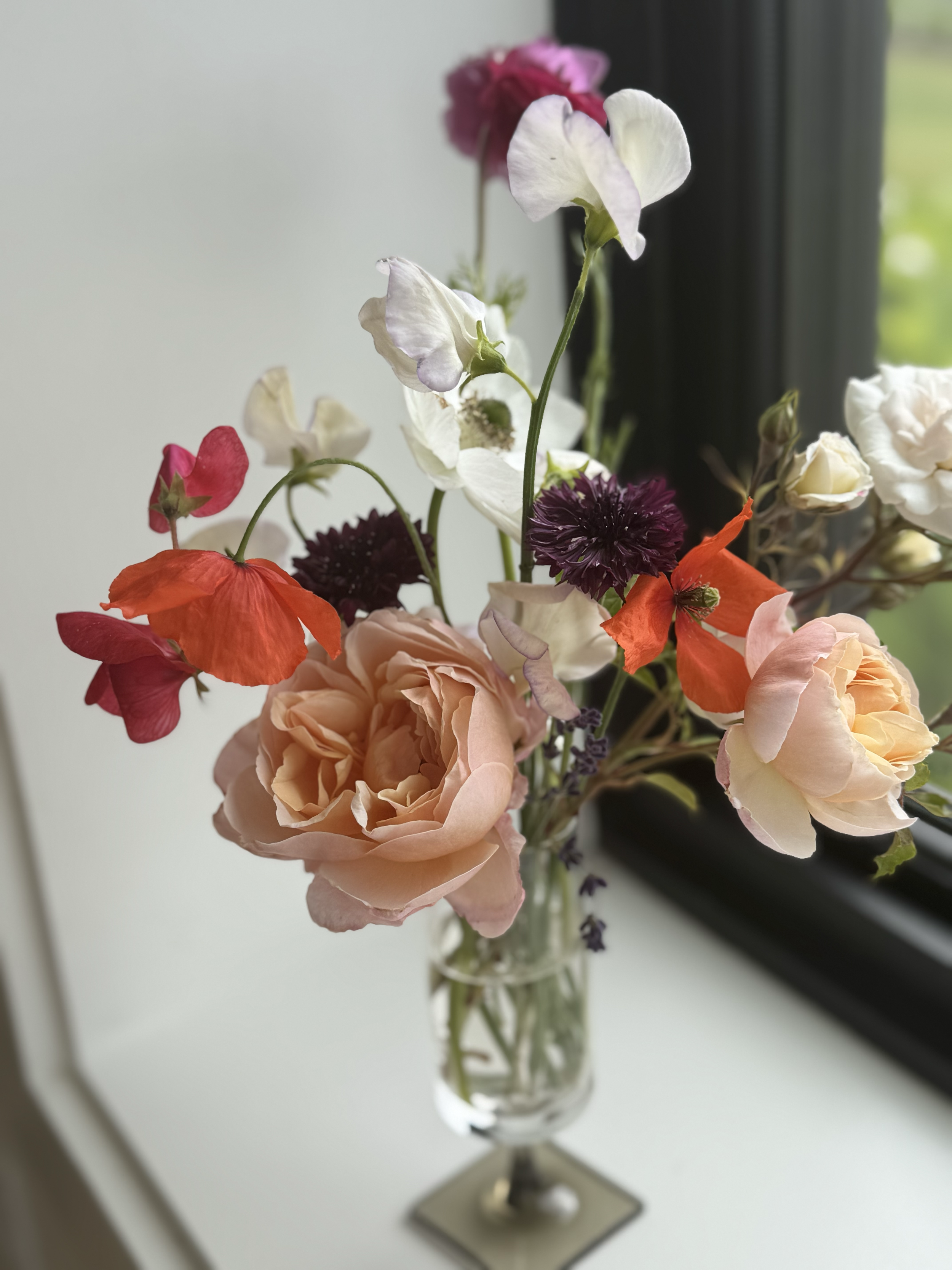 A vase with mixed pink, white, orange, and purple flowers on a white surface near a window.