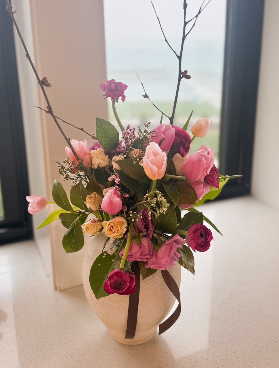 A vase with a bouquet of pink, purple, and yellow flowers, along with green leaves and branches, sitting on a white surface near a window.