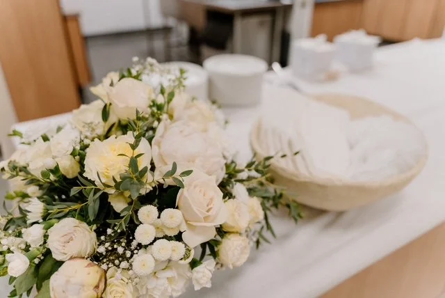 A bouquet of white and cream-colored flowers on a table, with a basket of napkins and some tableware in the background.