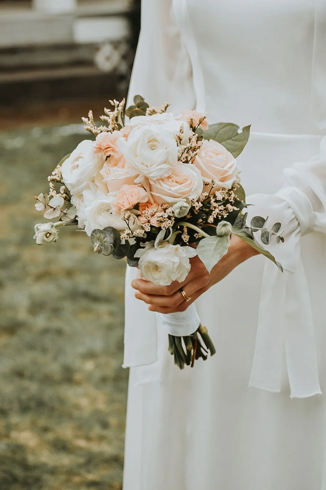A person in a white dress holding a small bouquet of white and blush roses with greenery.