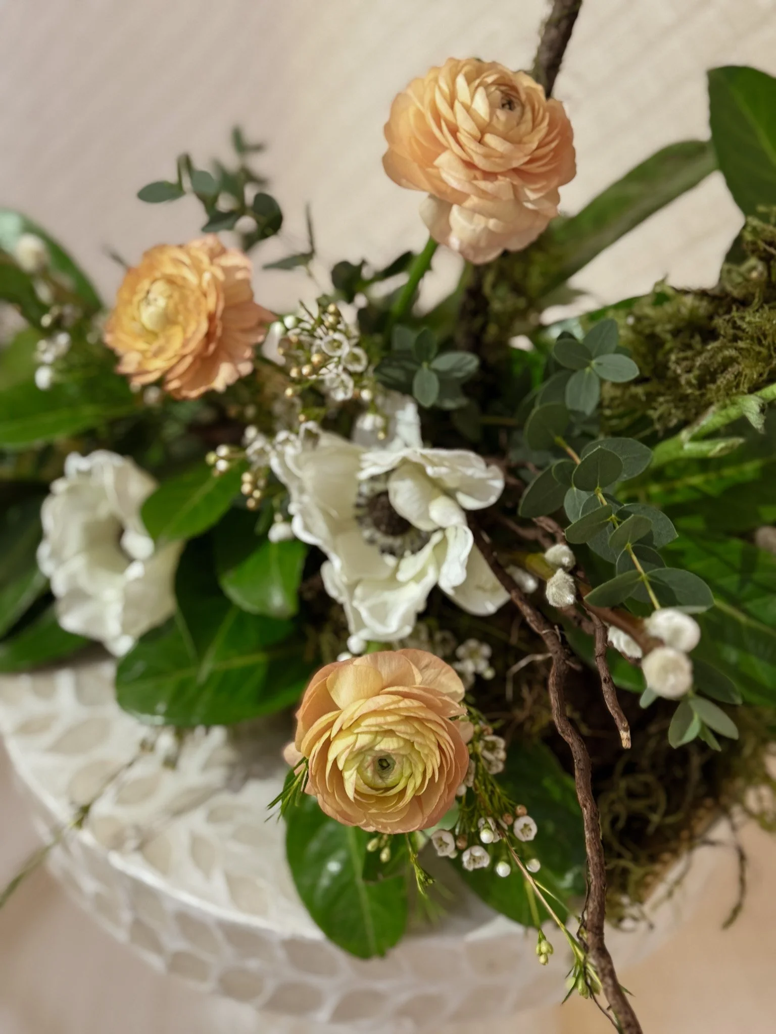 A floral arrangement with peach ranunculus, white anemone, white wax flowers, and greenery in a decorative vase.