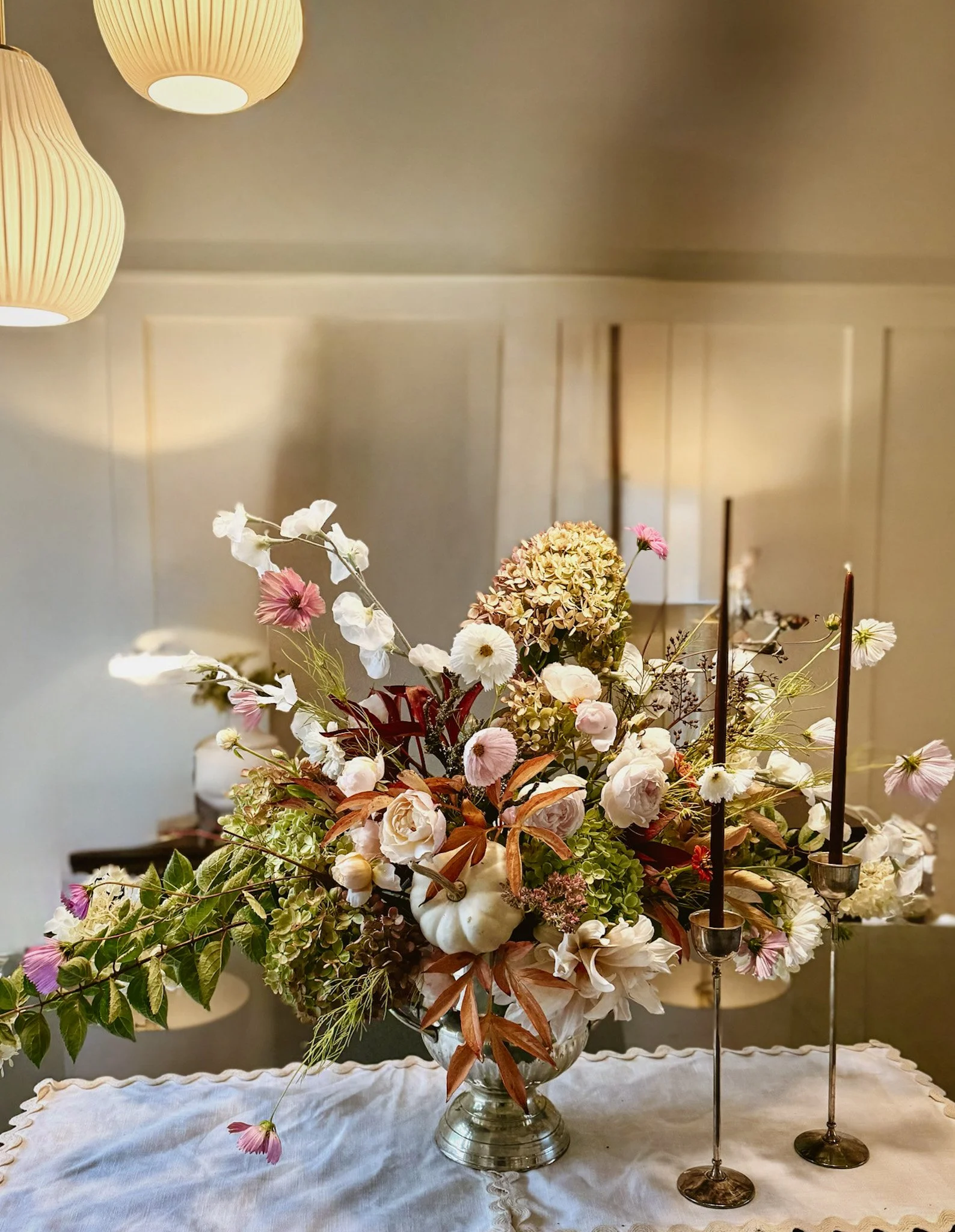 A floral centerpiece with white, pink, and green flowers and foliage, placed on a table with a white lace tablecloth, with two tall black candles in silver candlesticks on the right side.