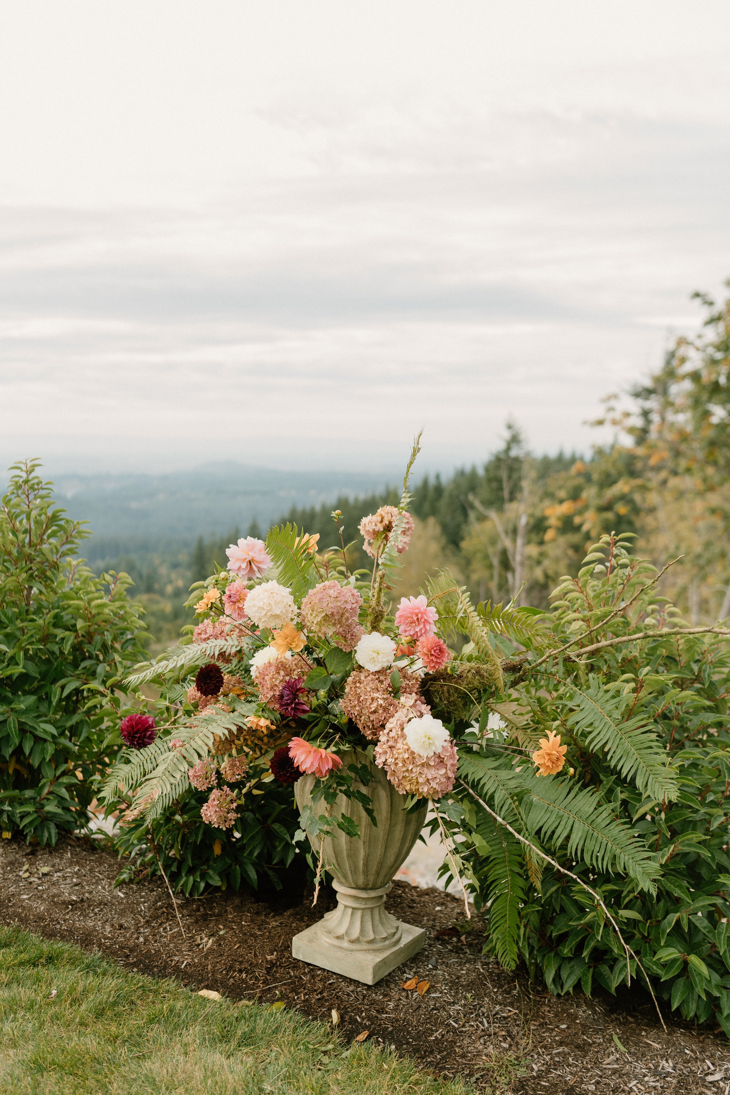 A large decorative stone urn filled with pink, white, yellow, and dark burgundy flowers, placed outdoors on a patch of soil, with green foliage and distant forested hills under a cloudy sky in the background.