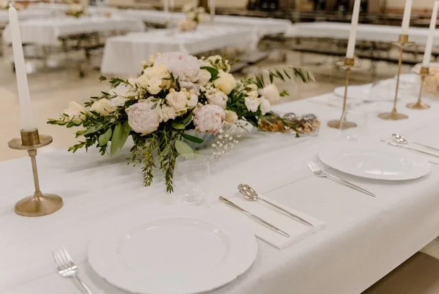 Elegant banquet table with white tablecloth, floral centerpiece with white and pink flowers, candles, and place settings for a formal event.
