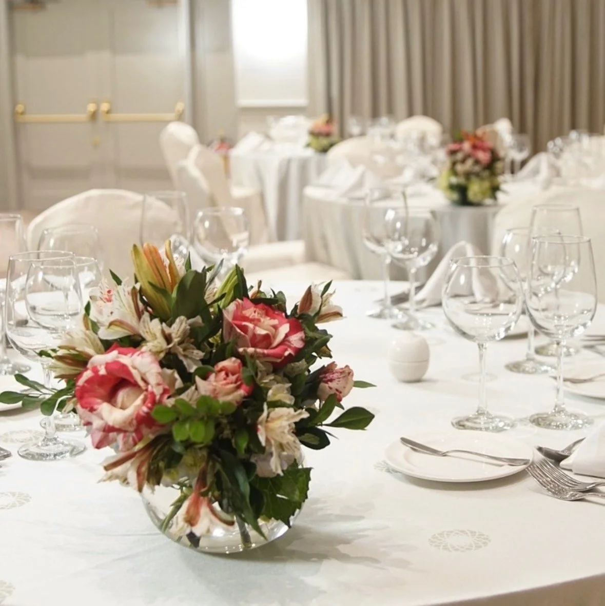 Elegant banquet table with floral centerpiece, multiple wine glasses, plates, and silverware in a well-lit room with gray curtains.
