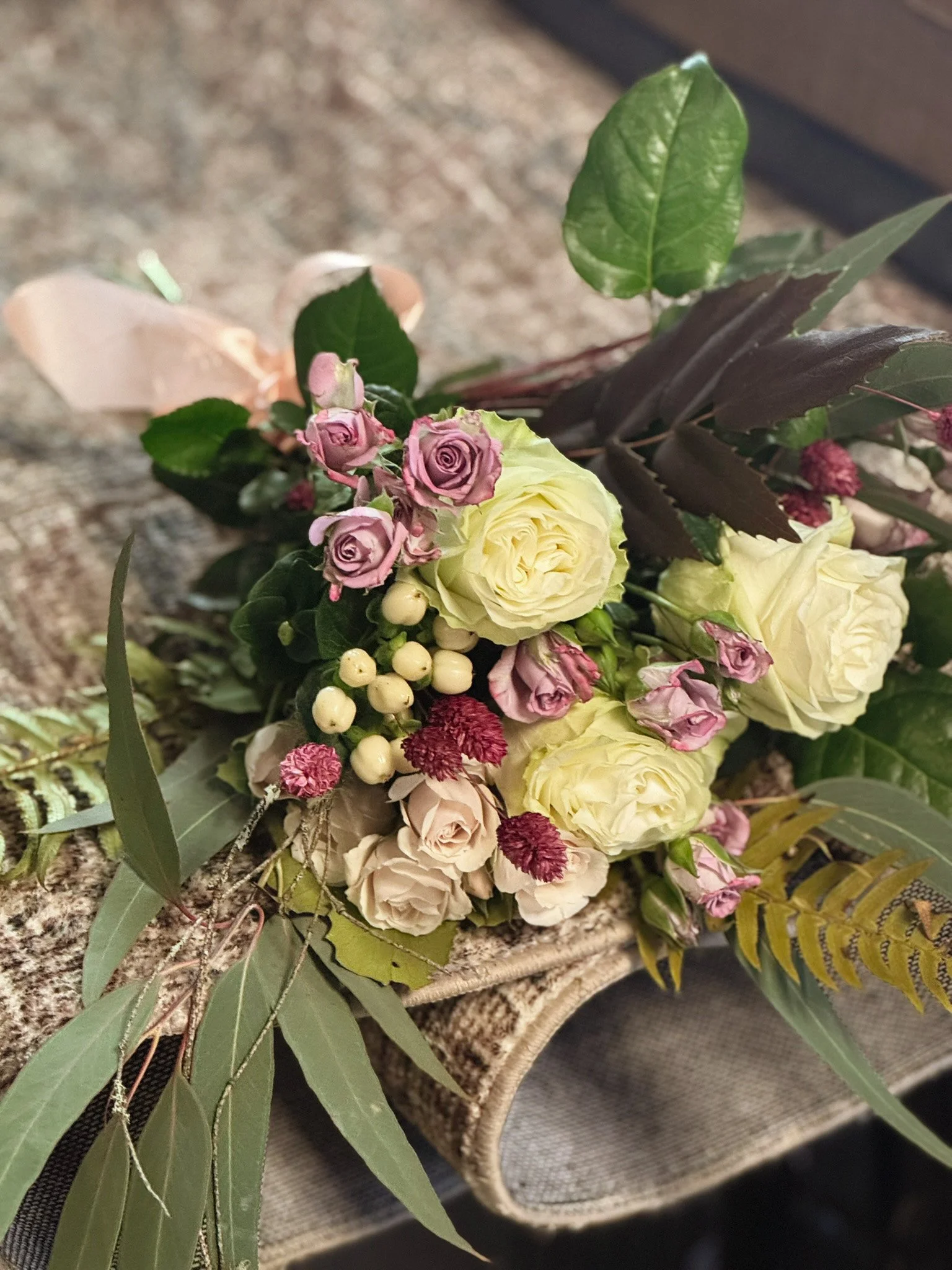 A small bouquet of mixed flowers including cream roses, purple roses, white berries, and red pom-pom flowers, arranged with green leaves on a textured surface.