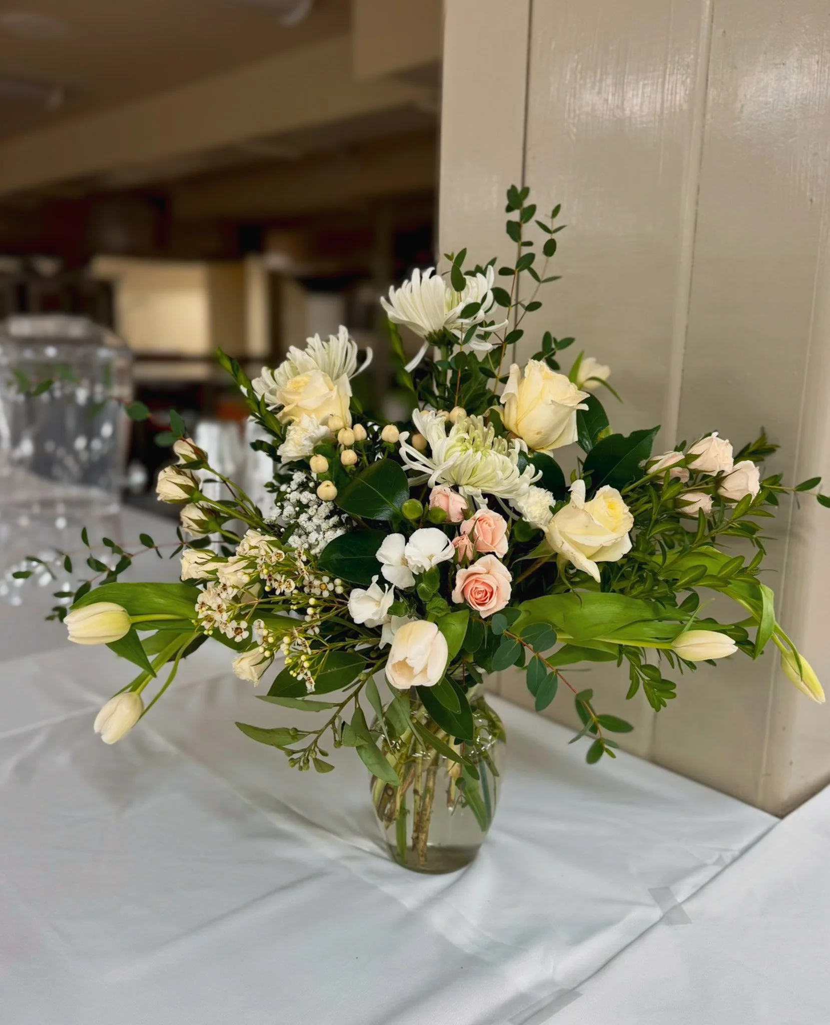 A floral arrangement with white and pale pink flowers and green foliage in a glass vase on a white tablecloth.