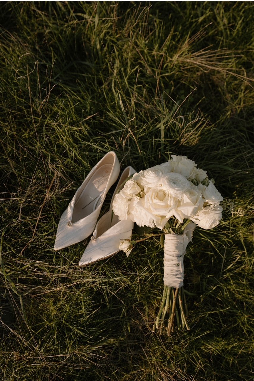 A pair of white high-heeled shoes and a bouquet of white flowers laying on grass.