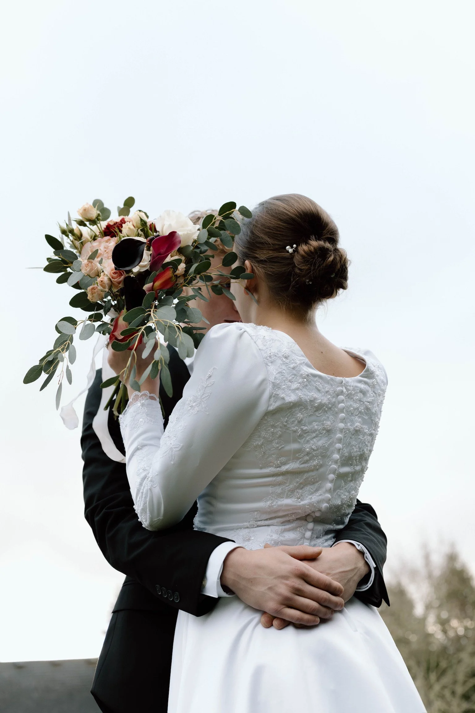 A bride and groom embrace outdoors on their wedding day, with the bride holding a bouquet of flowers.