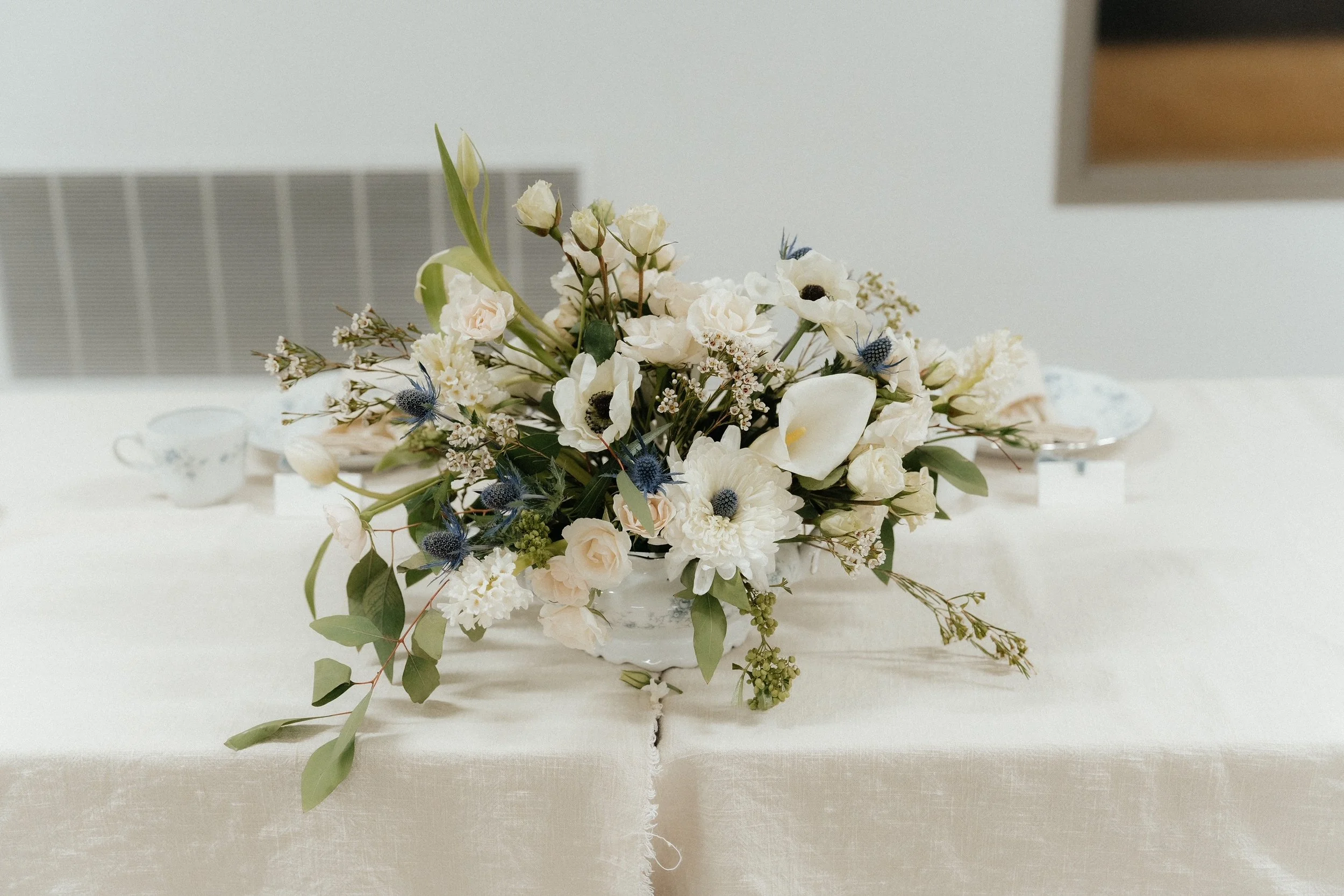 Elegant white flower centerpiece with green leaves on a beige tablecloth.