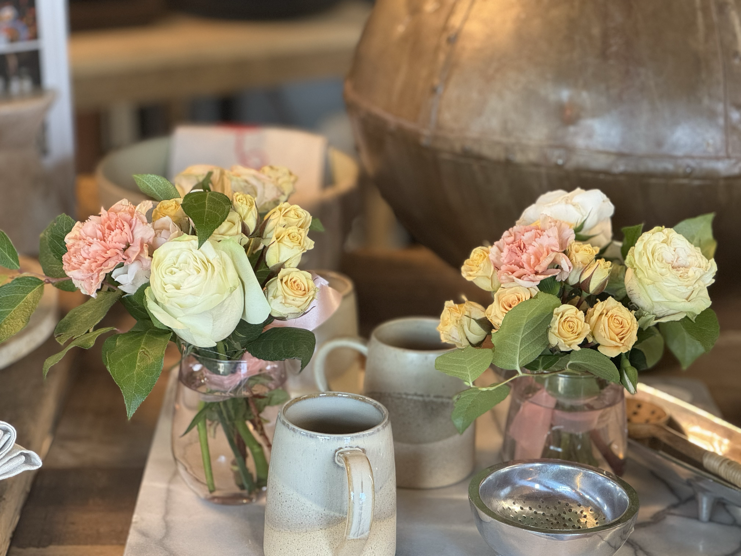 Vases with pink, white, and yellow roses on a wooden table, with ceramic mugs and a metal strainer nearby.