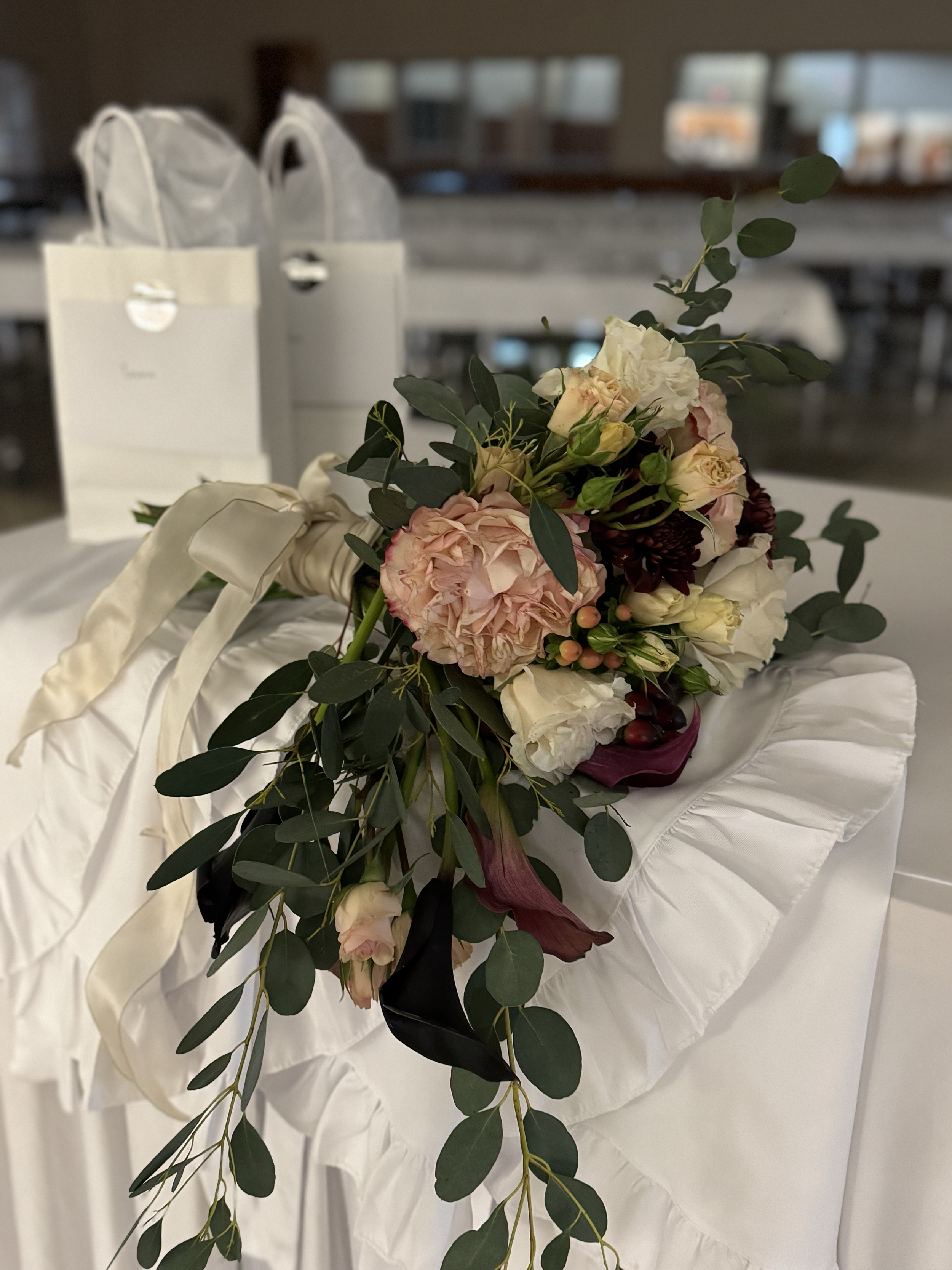 A bridal bouquet of pink, white, and dark red flowers with greenery, resting on a white ruffled cloth, with white gift bags in the background.