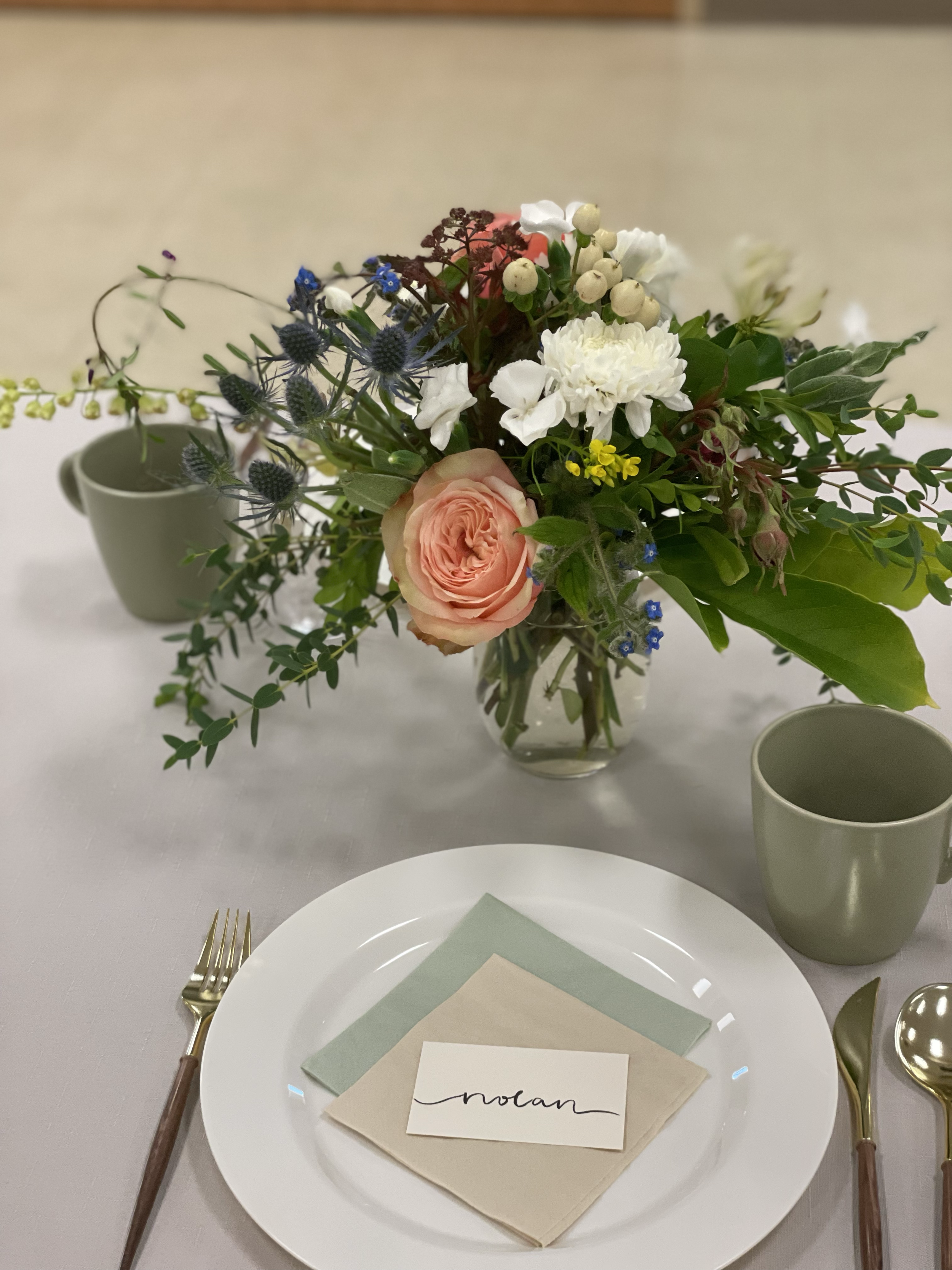 A table setting with a white plate, gold fork and spoon, a name card reading 'nolan', and layers of napkins, with a centerpiece of mixed flowers in a glass vase and two green cups.