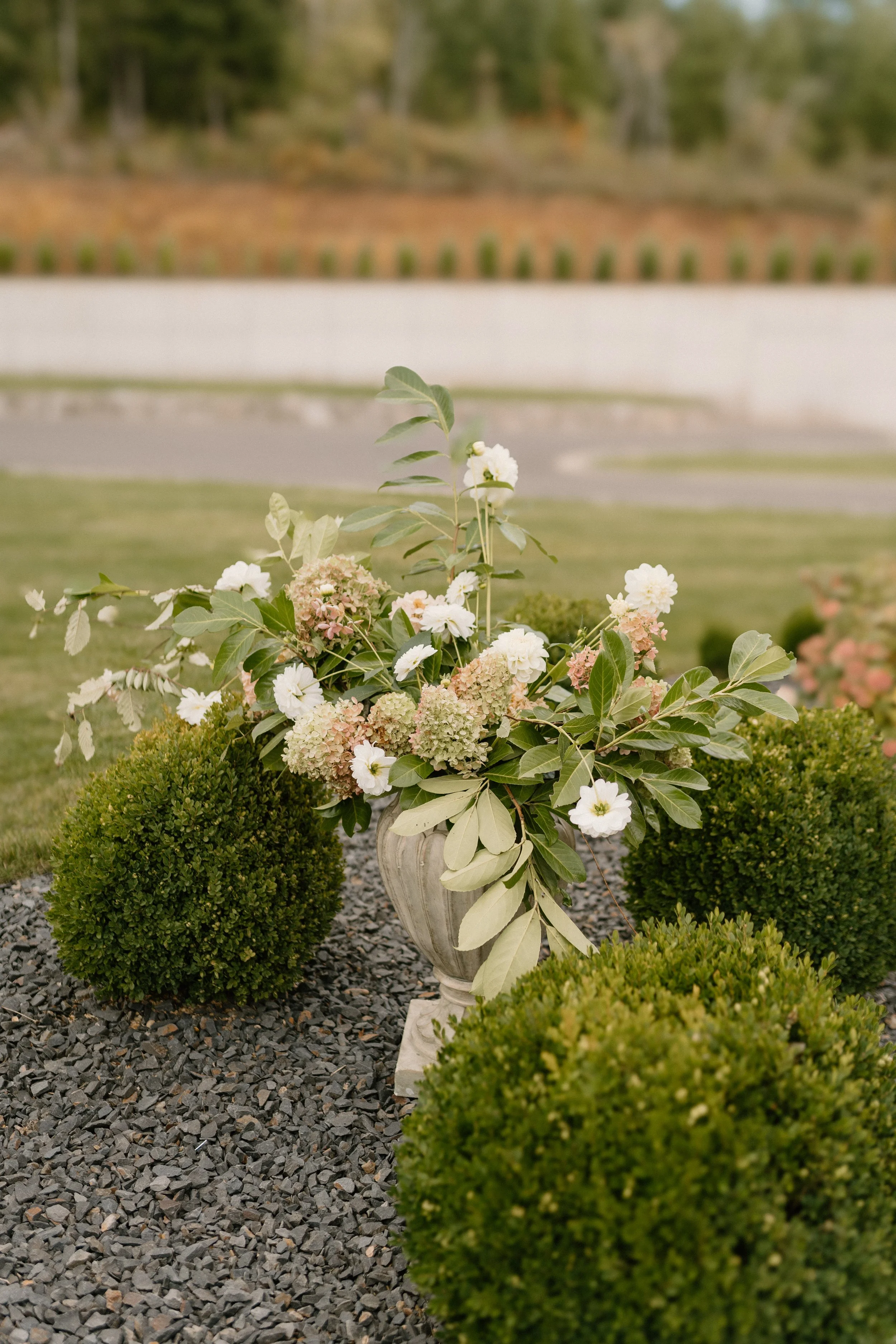 A floral arrangement in a decorative vase with green shrubbery around it, placed on a gravel surface in an outdoor garden setting.