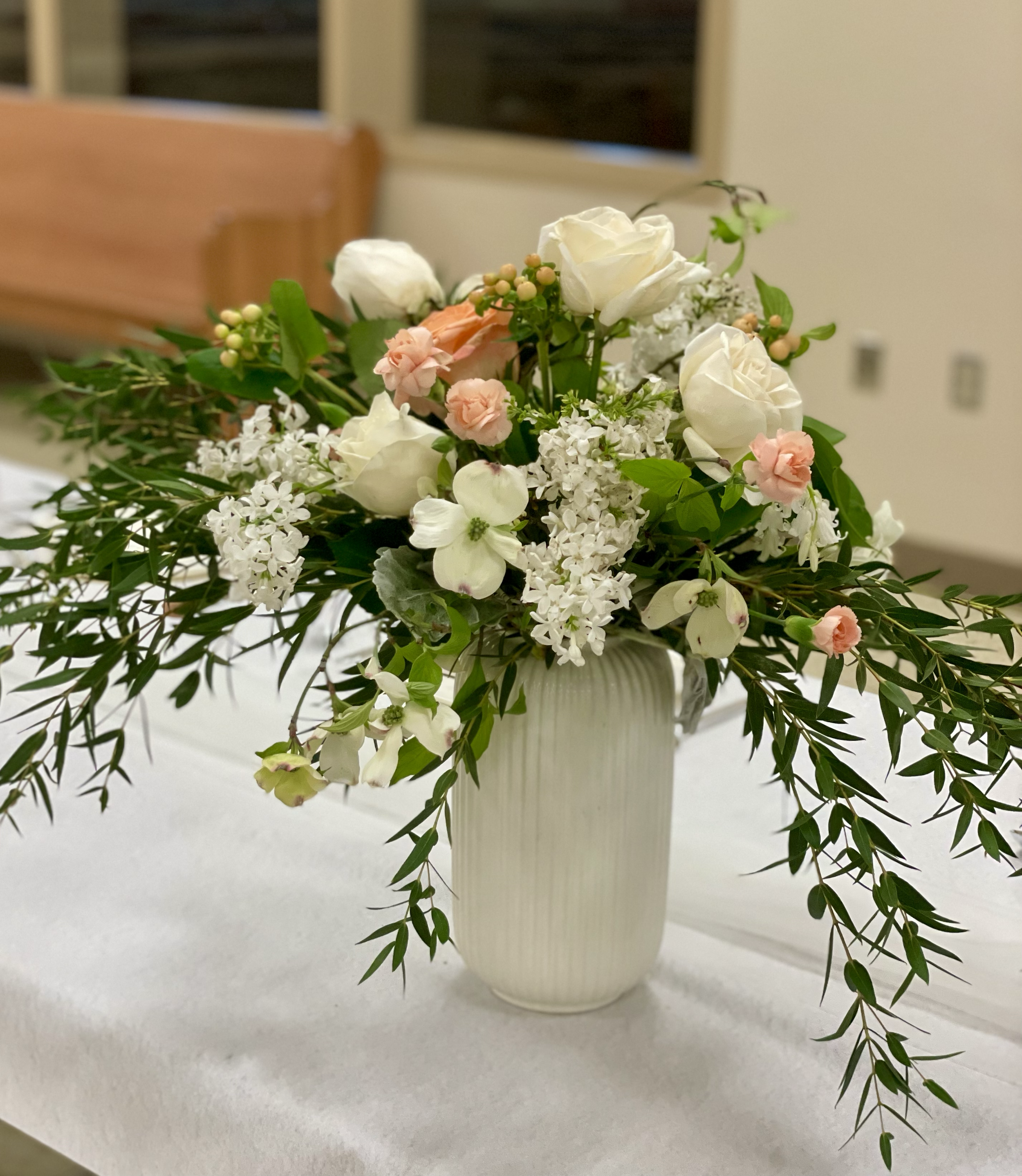A white vase with vertical ridges holding a mixed arrangement of white and light pink flowers, along with green foliage, placed on a white cloth surface.