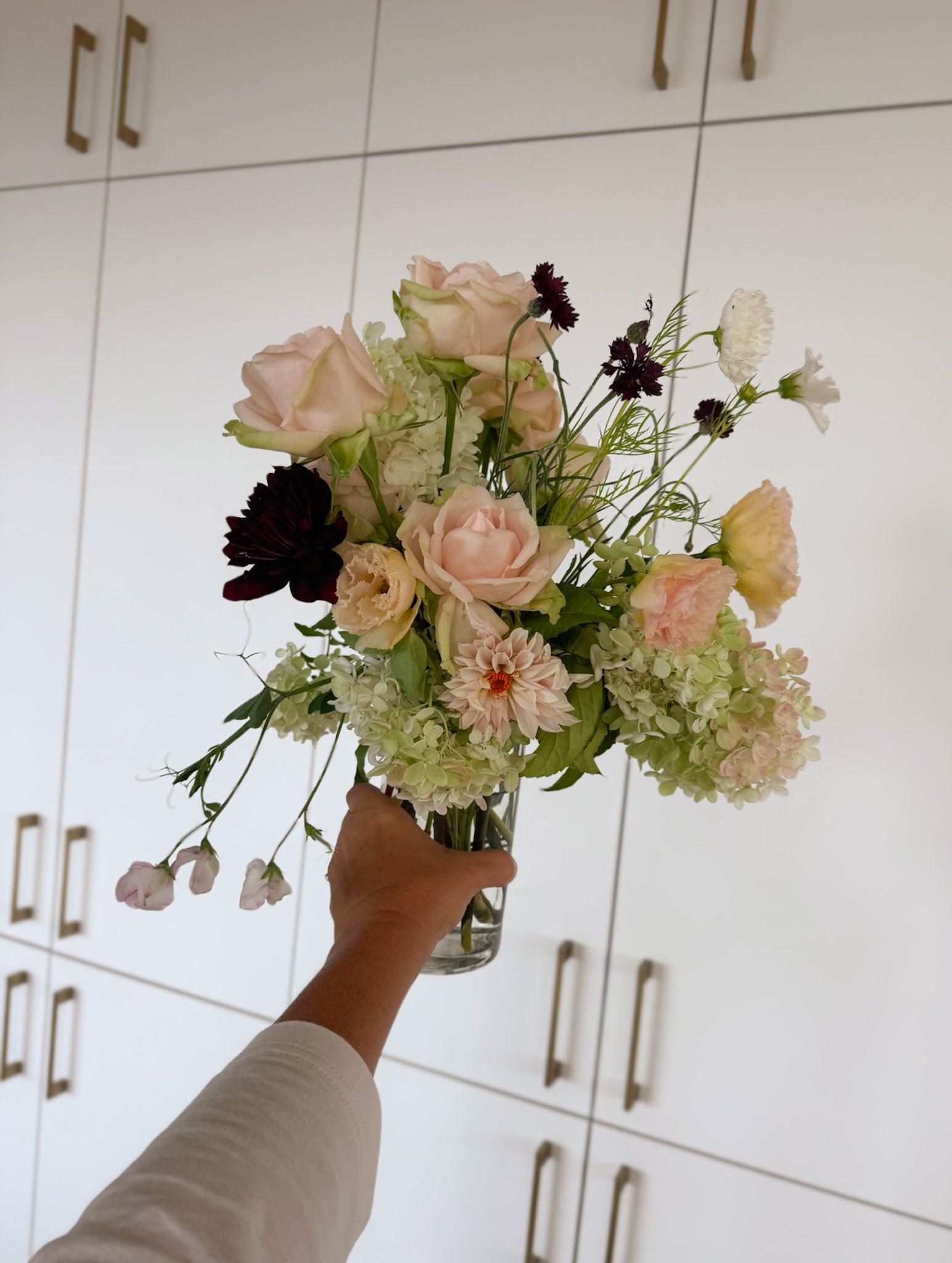 Hand holding a clear glass vase with a mixed bouquet of pink, cream, and dark purple flowers against white cabinets with gold handles.