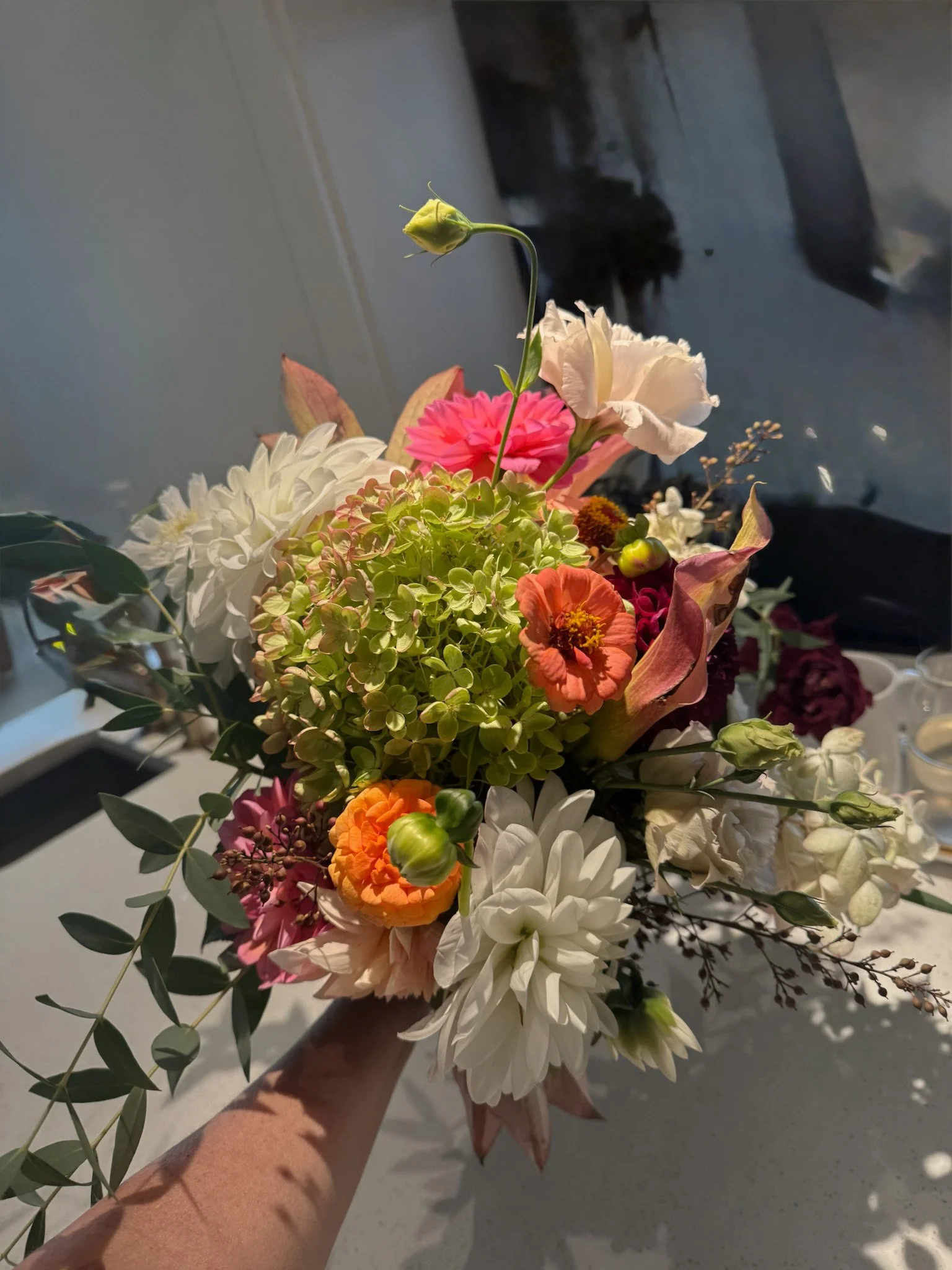 A person holding a colorful bouquet of mixed flowers including white, pink, orange, and purple blossoms, with some green foliage.